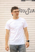 Man wearing a white t-shirt and glasses standing against a textured wall with graffiti.