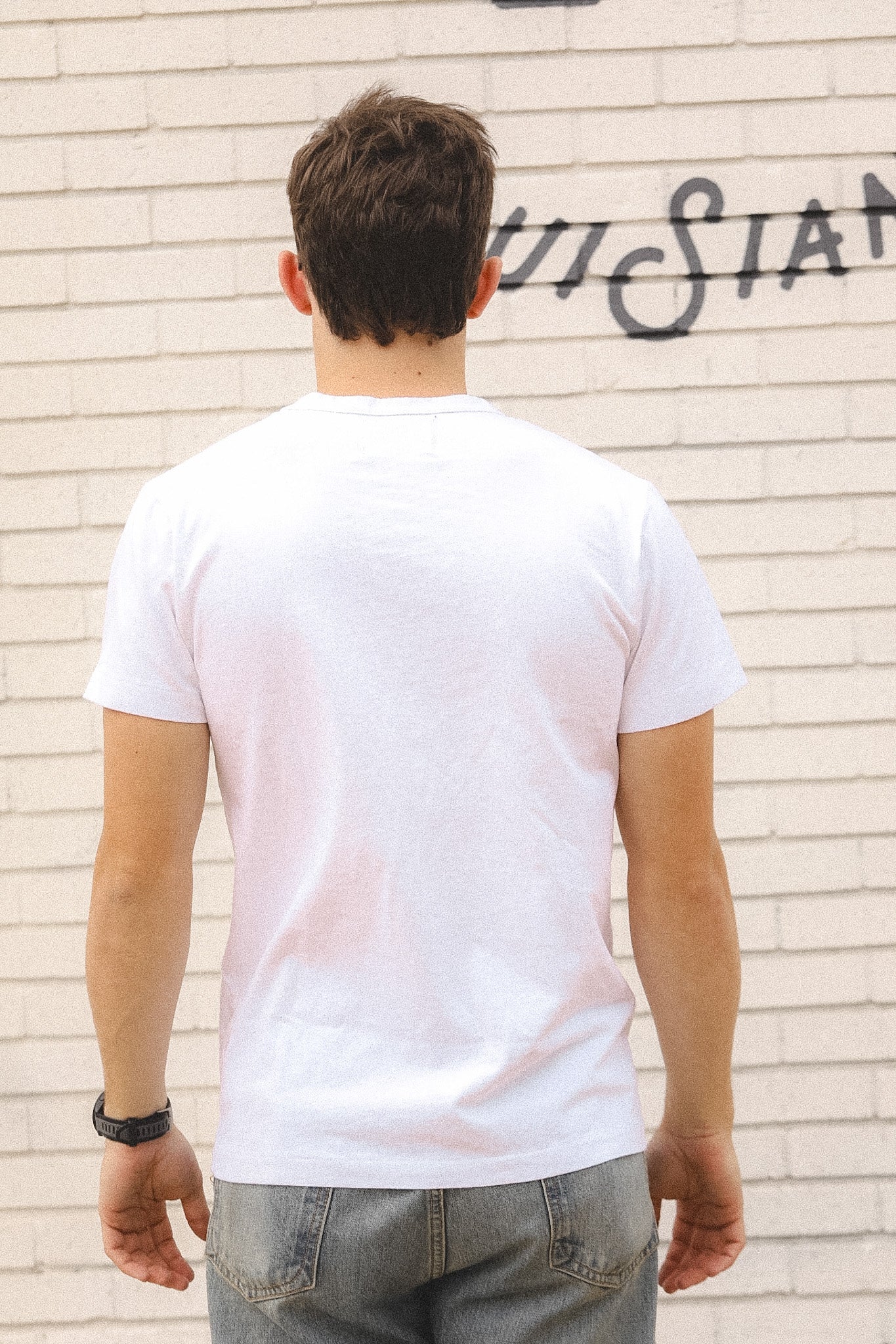 Person wearing a white t-shirt and jeans standing against a light brick wall with graffiti.