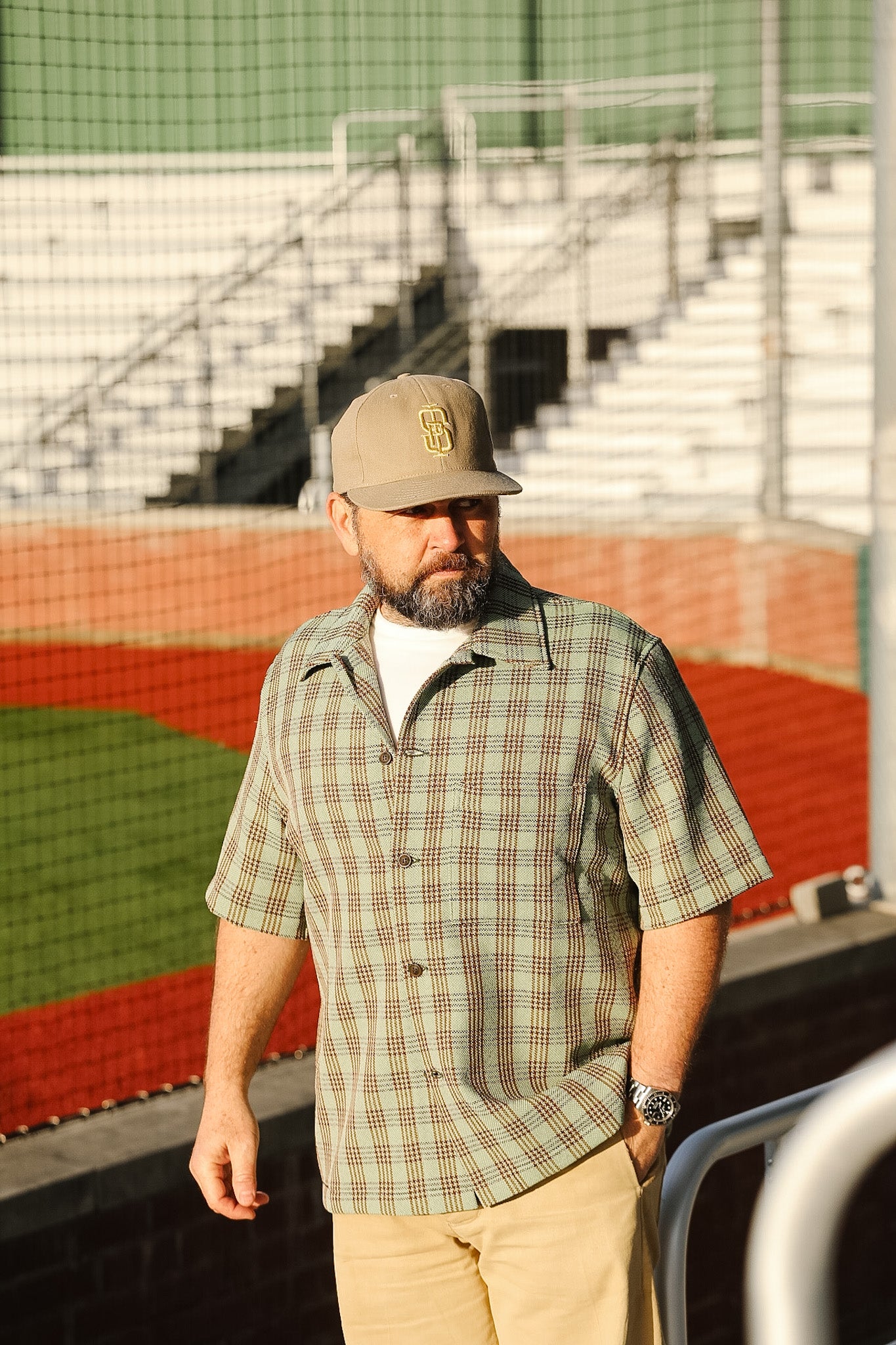 Man wearing a plaid shirt and cap standing in front of a sports field with bleachers.