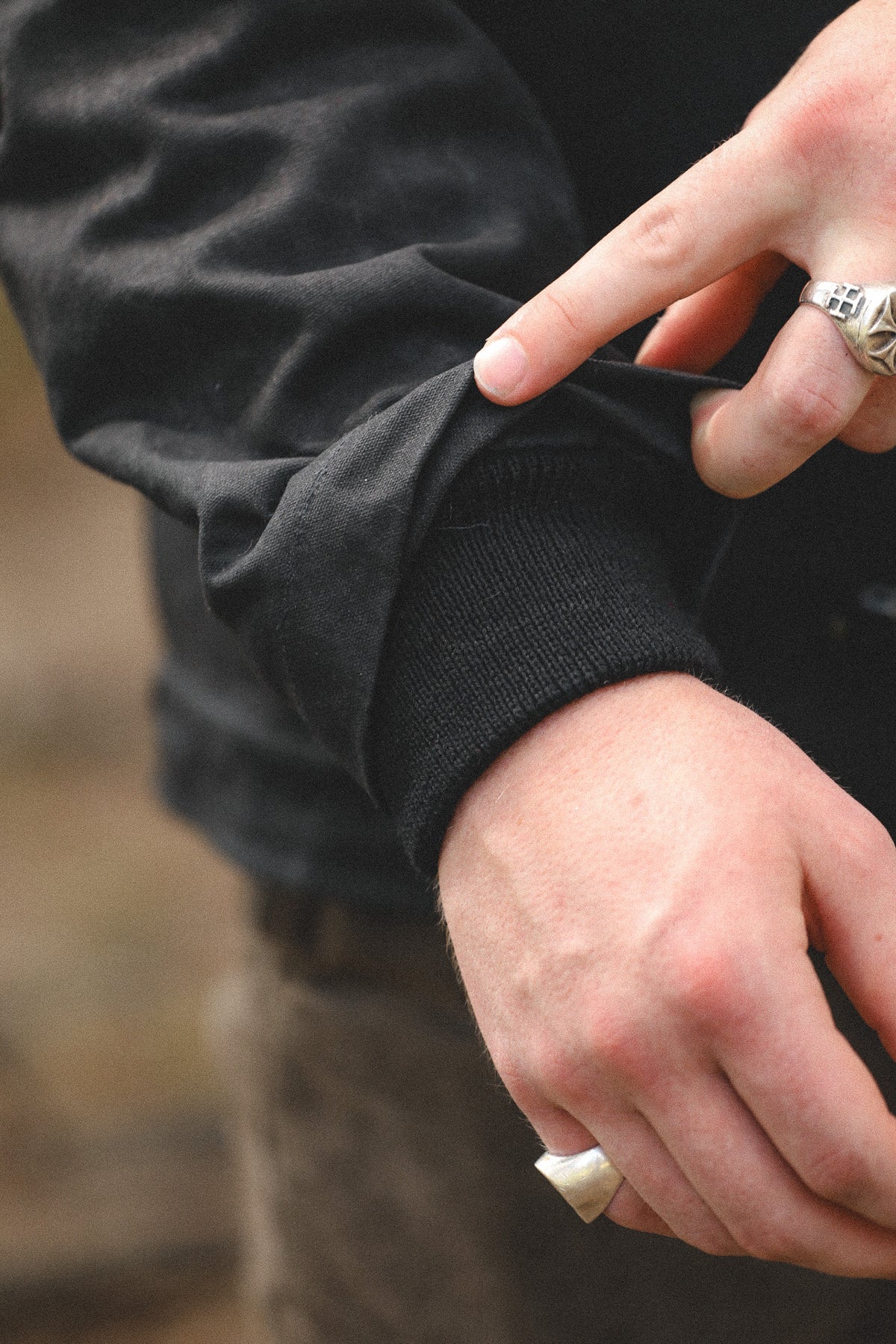 Close-up of a person wearing a black long-sleeve knit top, showing the ribbed cuff being pulled slightly by the hand, with silver rings on the fingers against a blurred outdoor background.