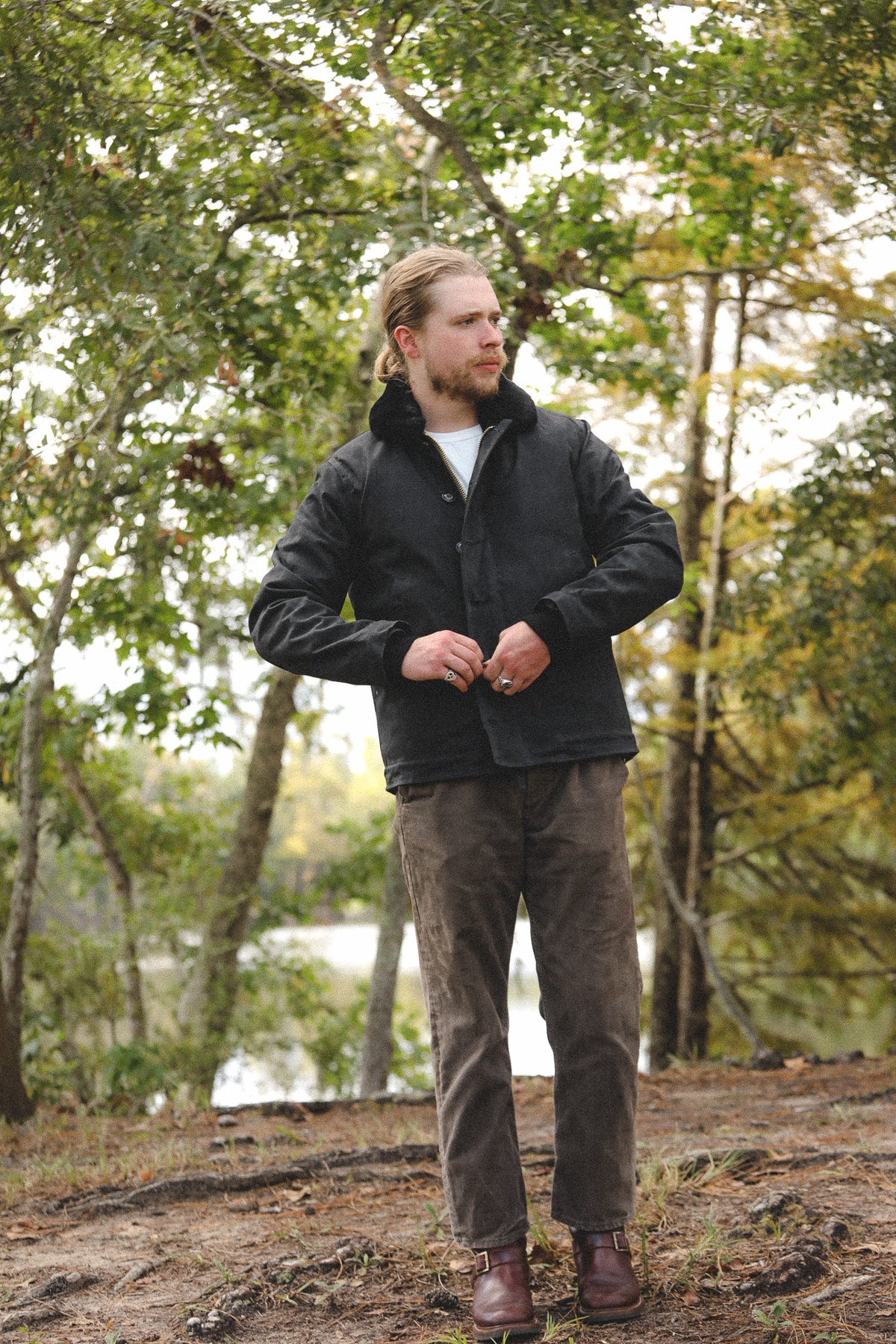 A man wearing a black zip-up jacket, brown pants, and brown boots standing outdoors in a wooded area near water.
