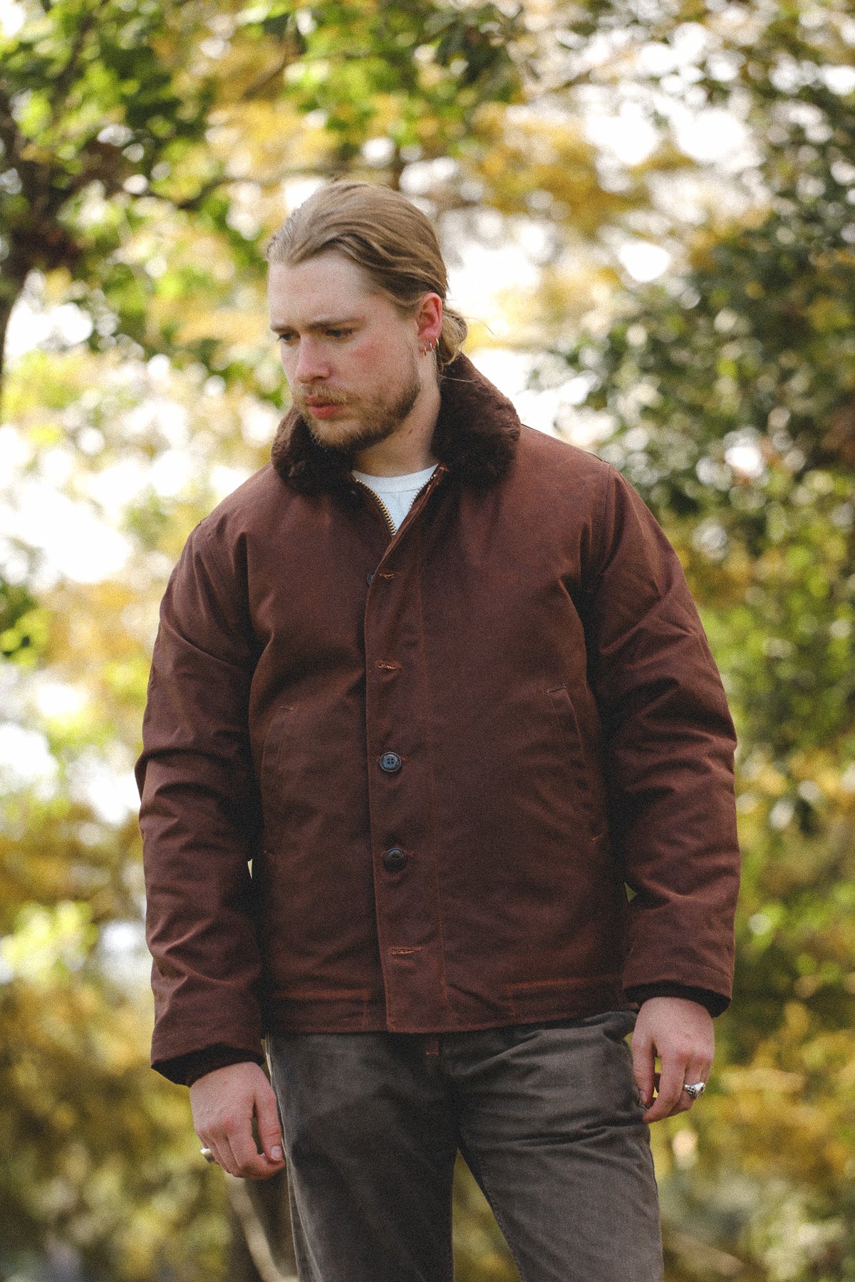 A man wearing a brown waxed cotton field jacket with a dark brown fur collar, standing outdoors against a blurred background of green and yellow autumn trees.