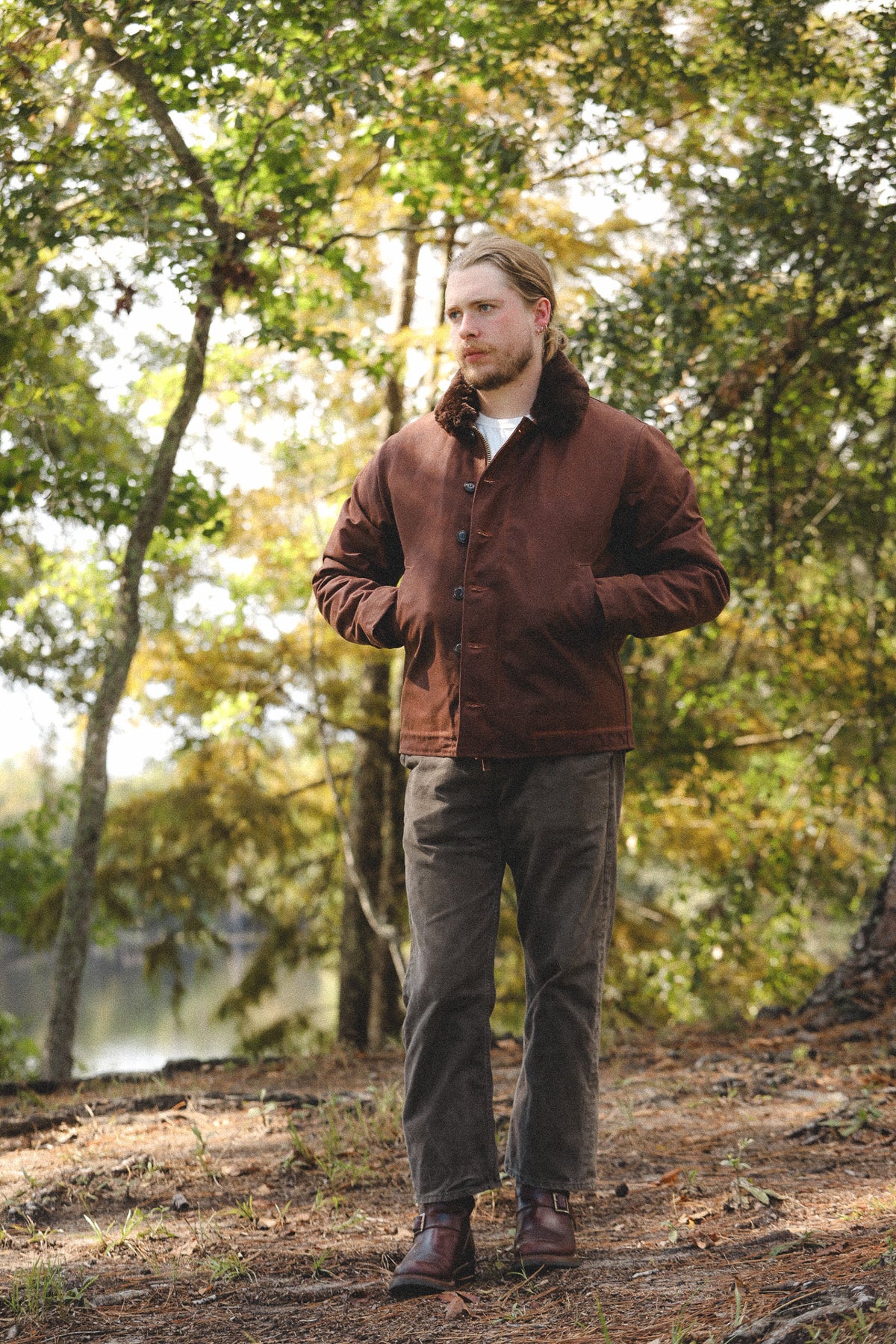 A man wearing a brown corduroy jacket with a dark brown fur collar, gray pants, and brown boots stands in a wooded outdoor setting.