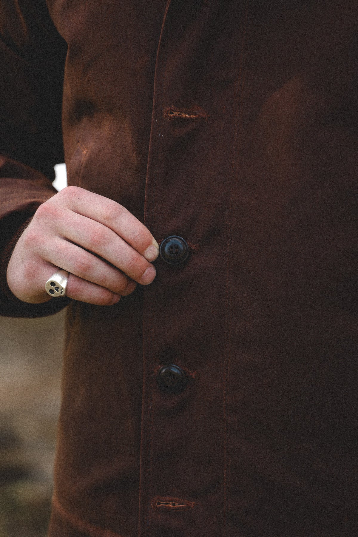 Close-up of a person wearing a dark brown waxed cotton jacket, with their hand adjusting a dark button. A silver ring is visible on their finger.