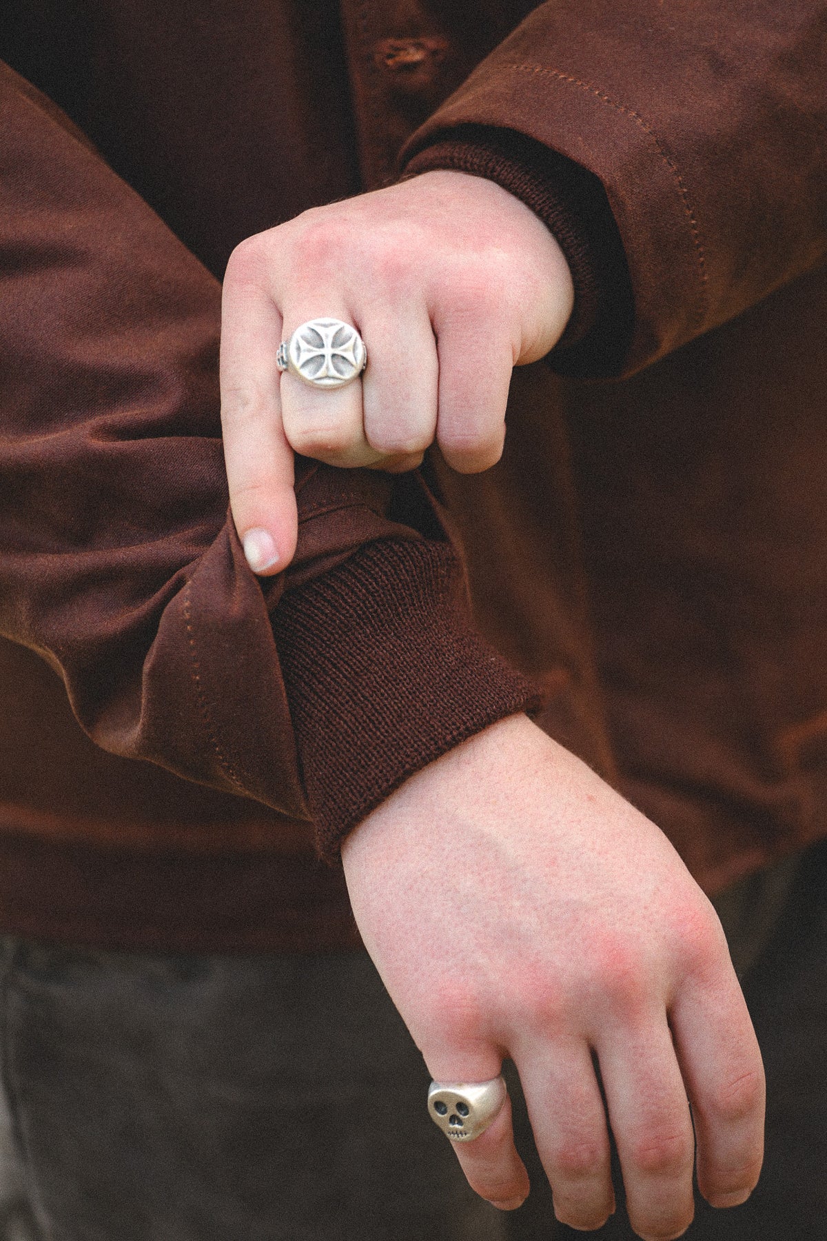 A close-up of a person wearing silver-toned signet rings, featuring a circular ring with a cross-like emblem on one finger and a smaller matching ring on another finger, set against a brown jacket.