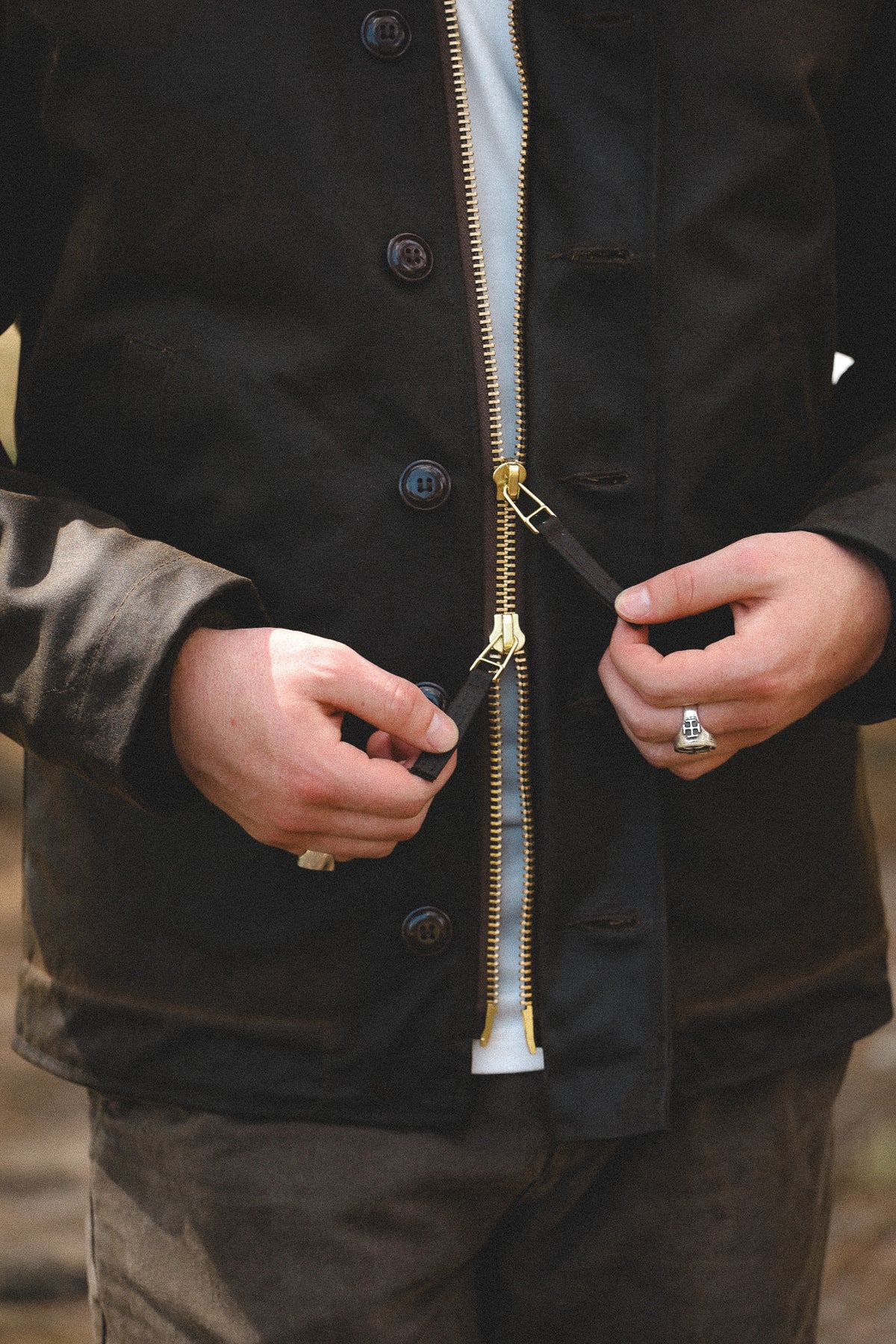 Close-up of a person wearing a black waxed canvas jacket with a gold zipper, opening it to reveal a white shirt underneath.