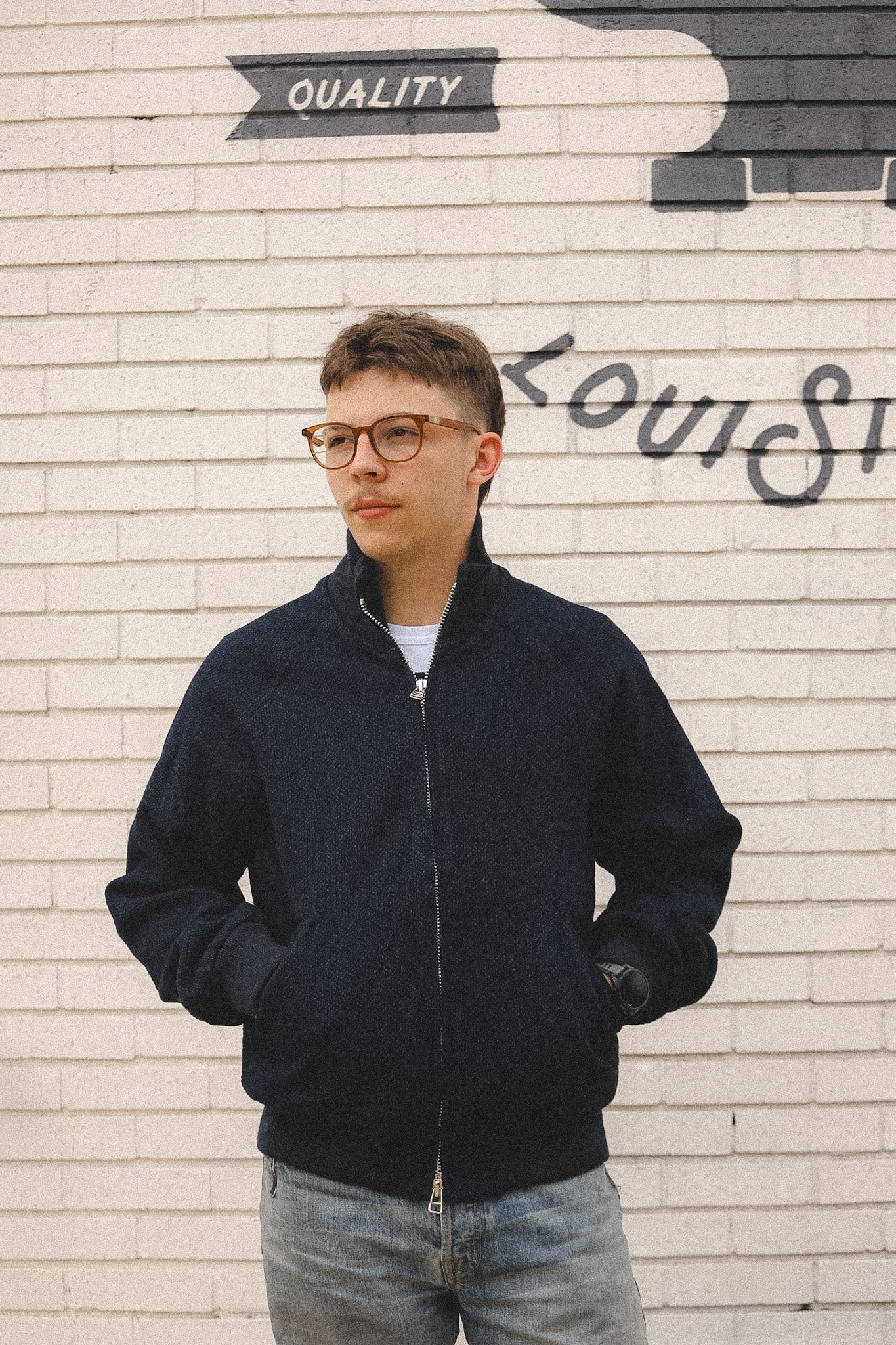 Person wearing a navy bomber jacket standing in front of a textured wall with graffiti.