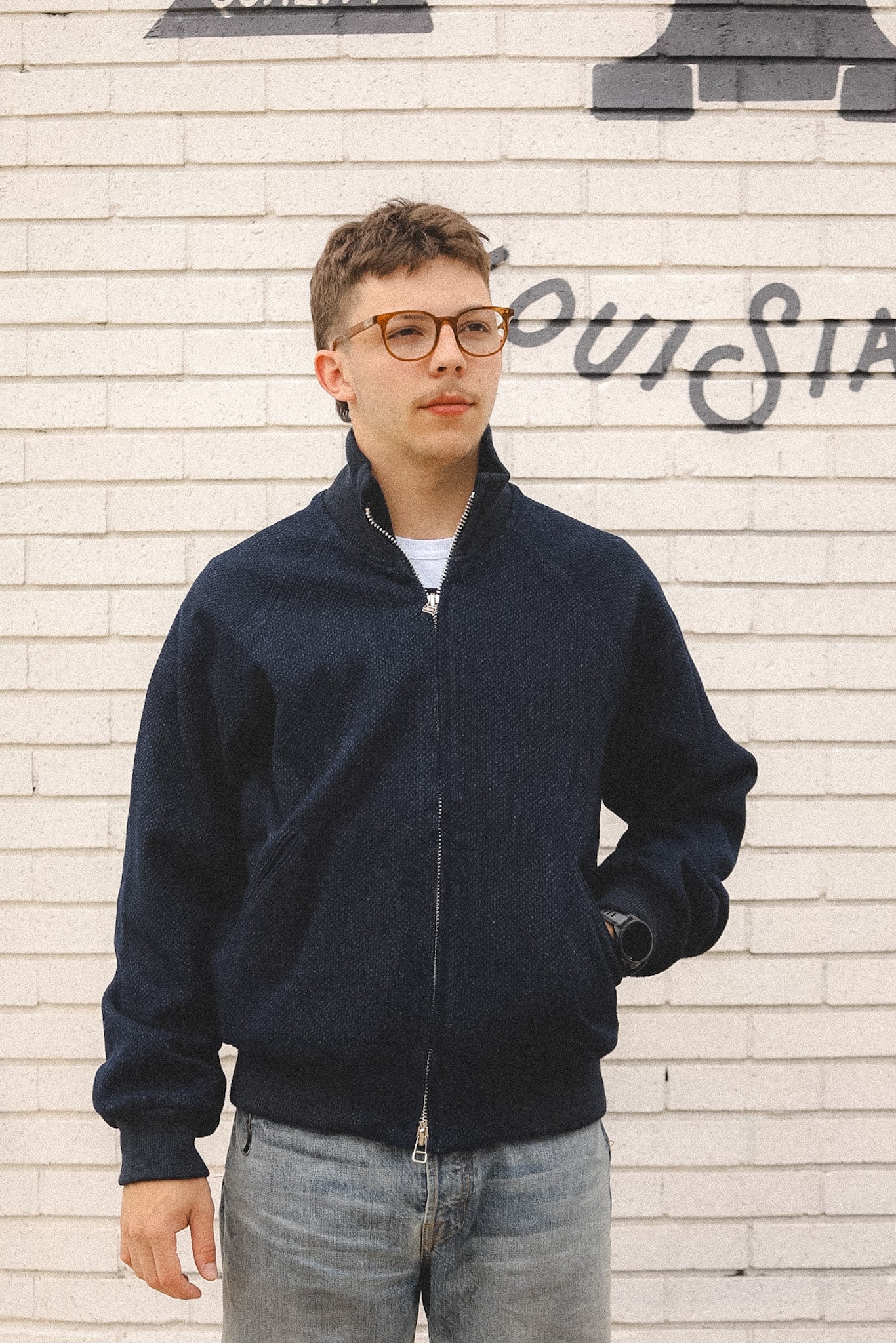 Man wearing a navy bomber jacket and glasses standing against a textured wall.