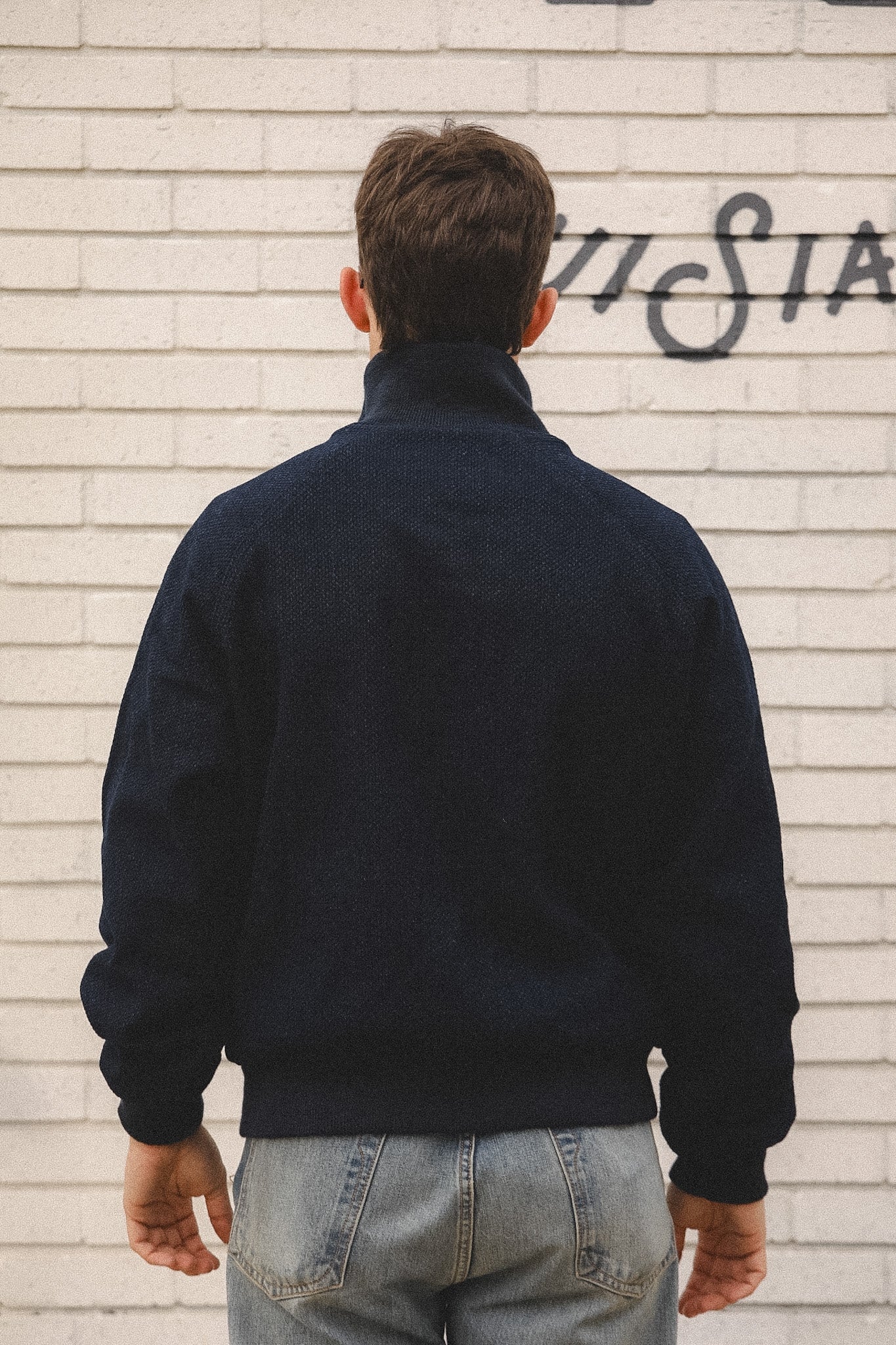 Person wearing a dark jacket and light blue jeans standing against a textured wall with graffiti.