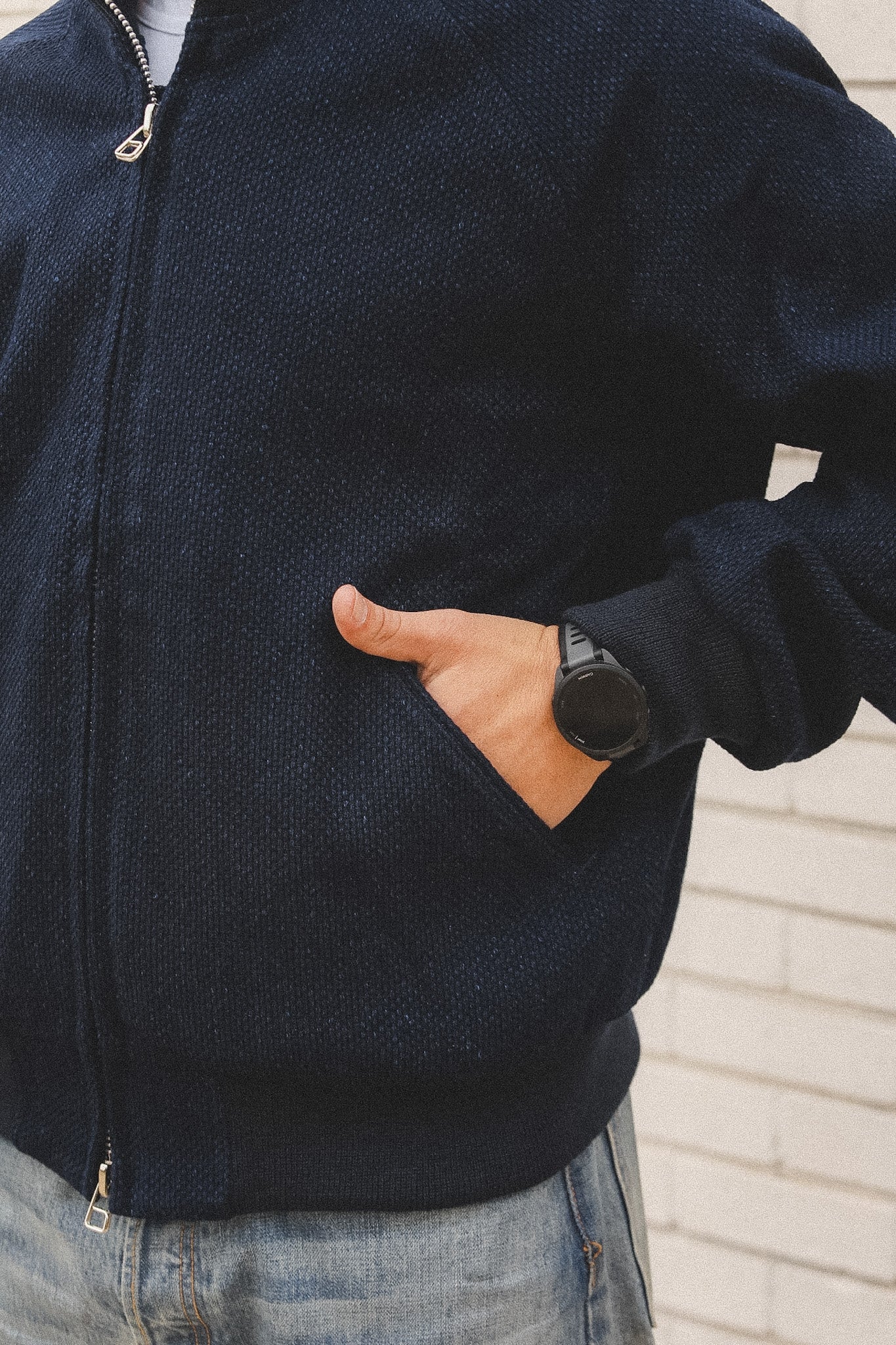 Person wearing a dark blue jacket with a hand in the pocket, standing against a light-colored wall.
