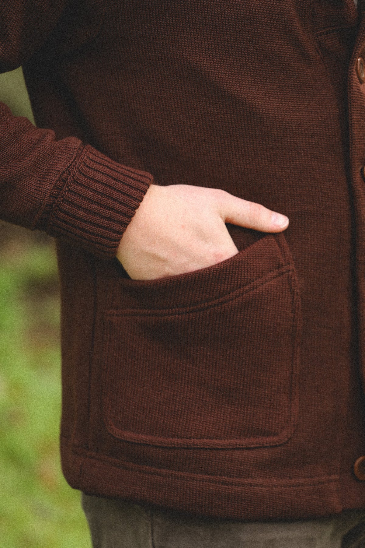 Close-up of a person wearing a dark brown ribbed knit cardigan with a front patch pocket and a hand tucked into the pocket.