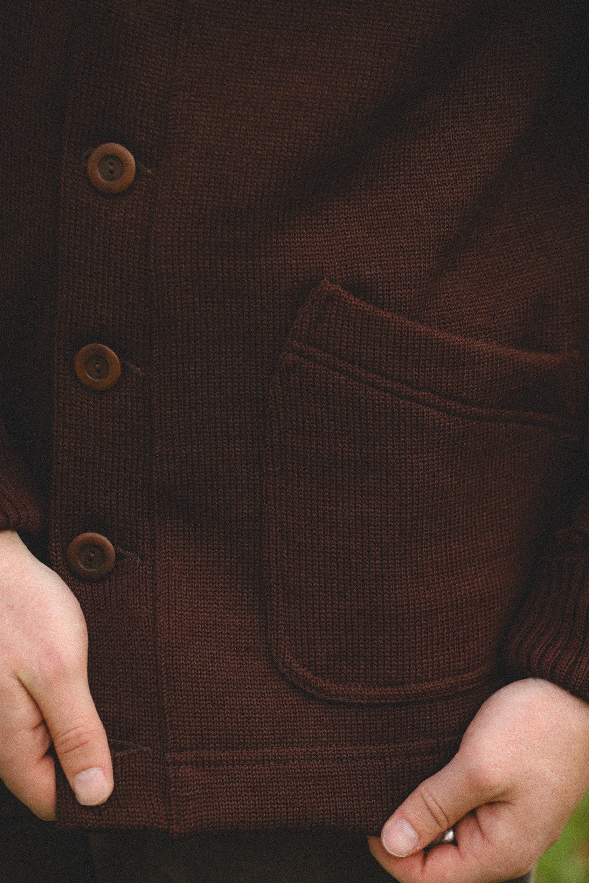 Close-up of a person wearing a dark brown ribbed knit cardigan sweater with three brown buttons and a front patch pocket, with hands holding the hem.