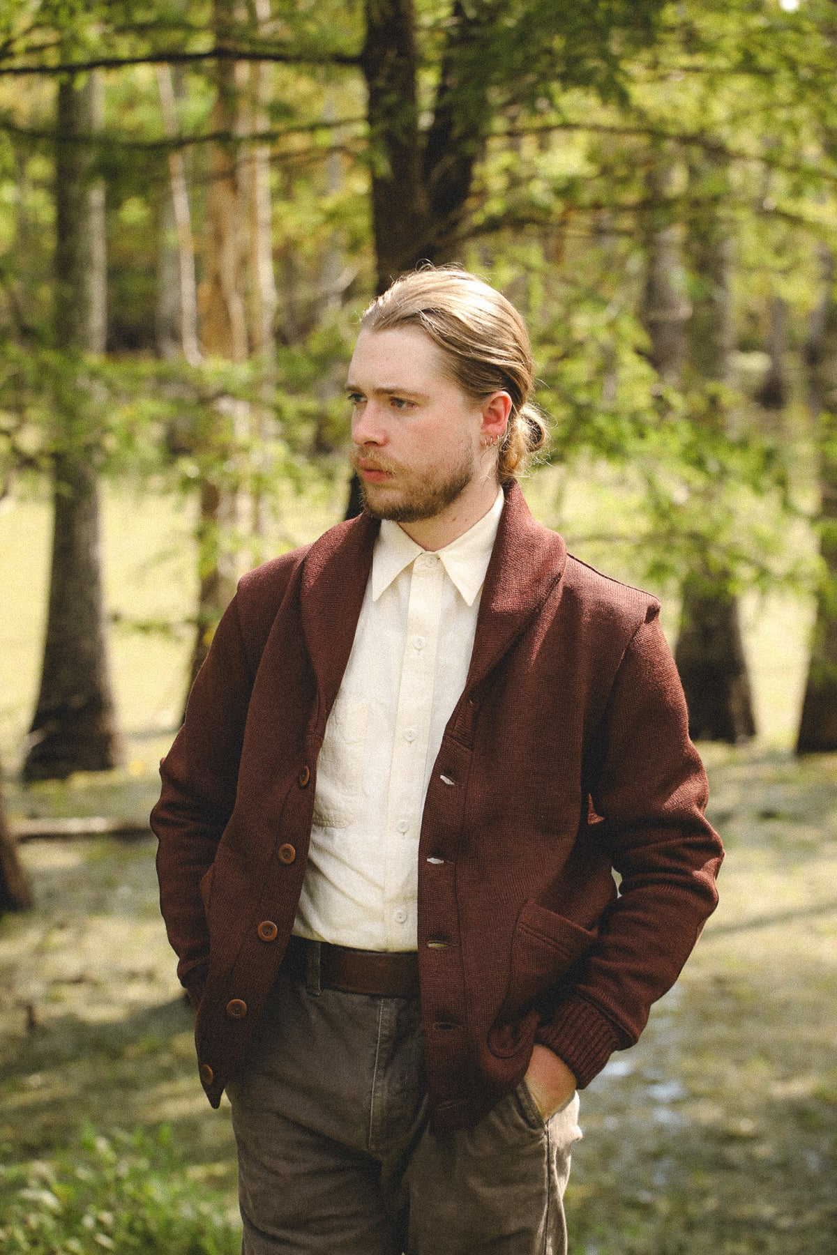 A man standing in a sunlit forest wearing a dark brown cardigan over a cream shirt, with gray trousers and his hands in his pockets.