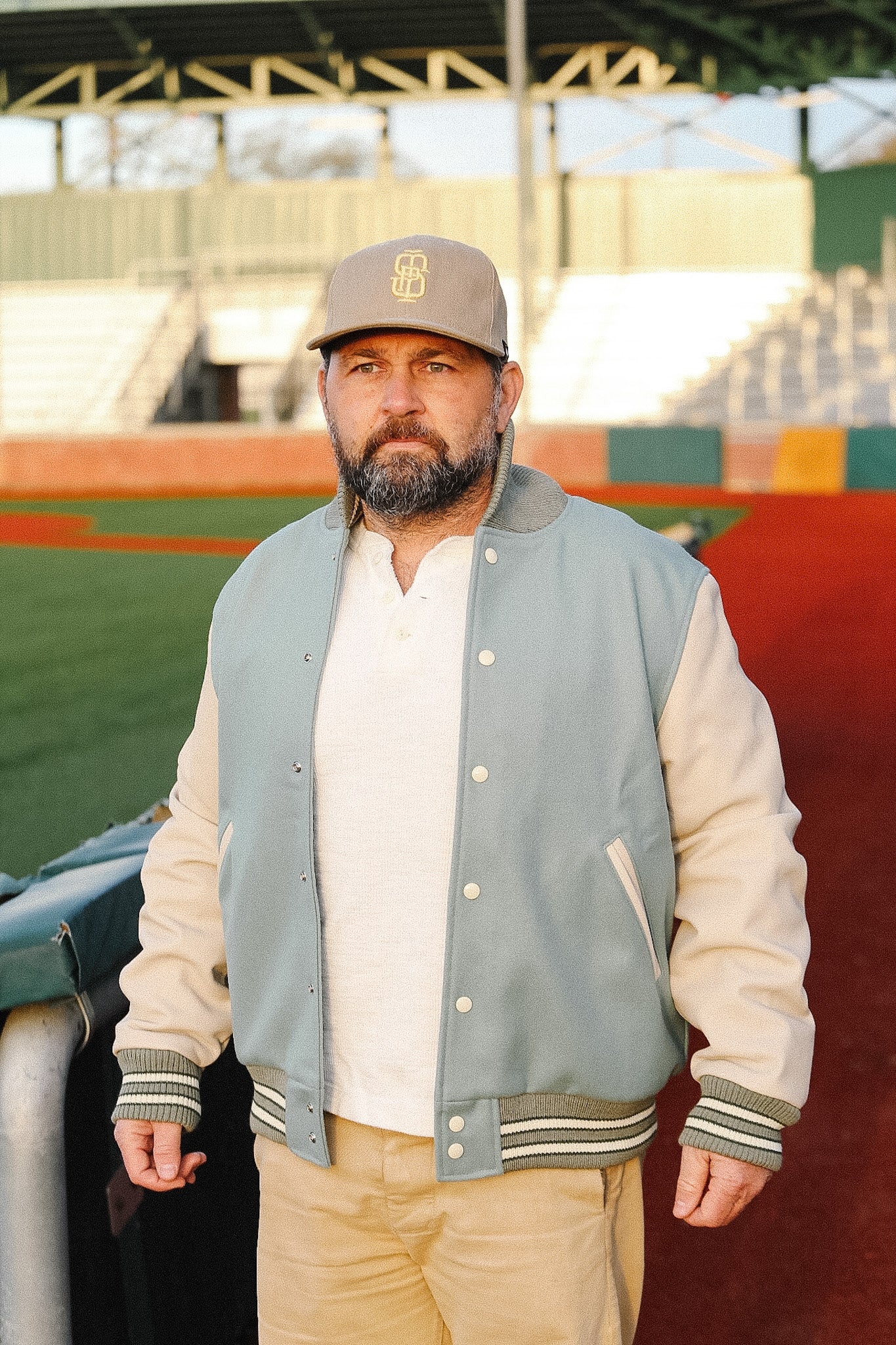 Man wearing a letterman jacket and cap standing on a sports field.