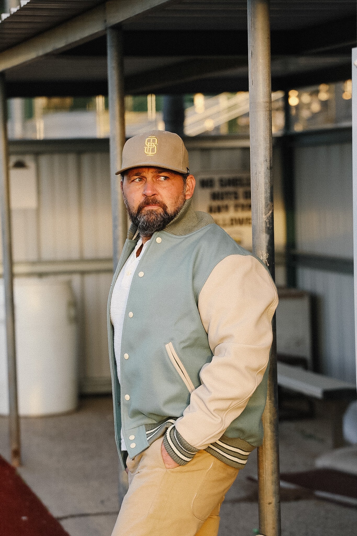 Man wearing a varsity jacket and cap standing in an industrial setting
