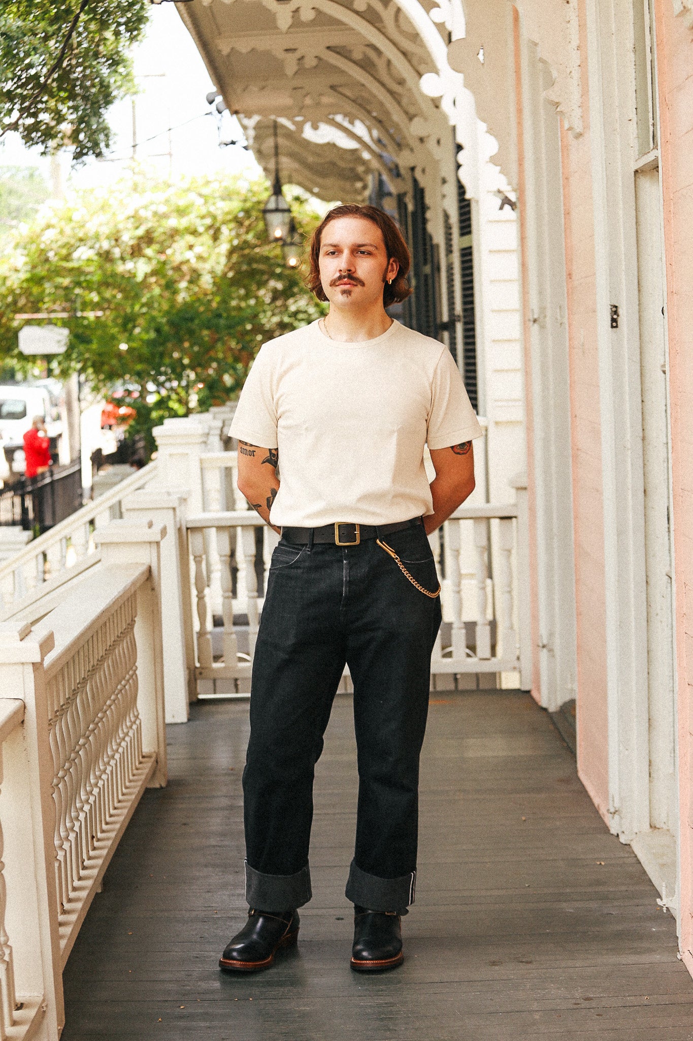 A man stands on a porch wearing a cream crewneck t-shirt, dark blue jeans with rolled cuffs, a brown leather belt with a gold buckle, and black leather shoes.