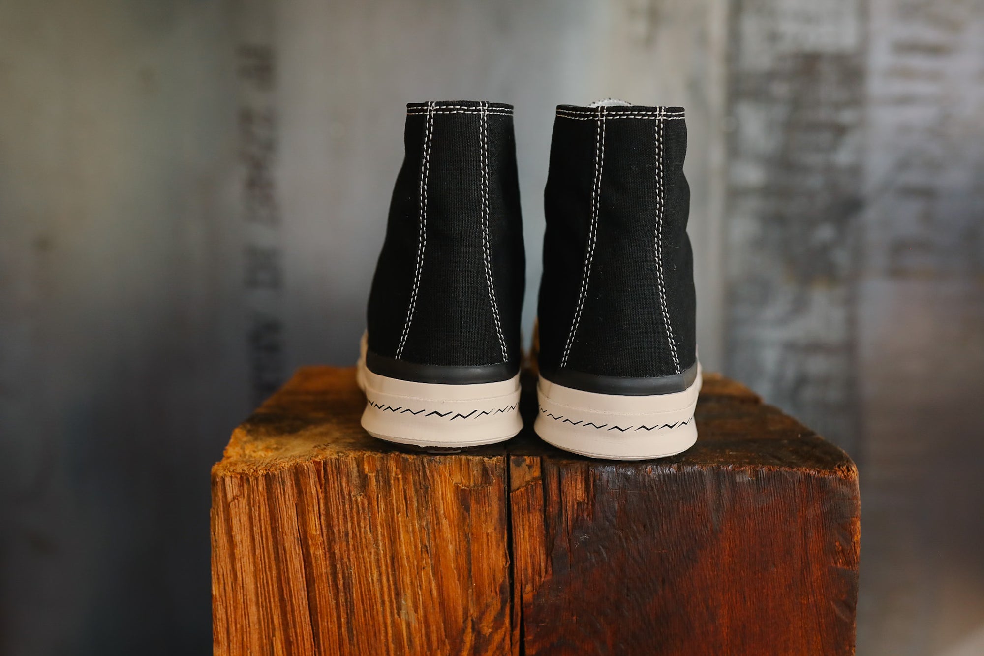 Pair of black high-top sneakers with white soles on a wooden block against a textured wall.