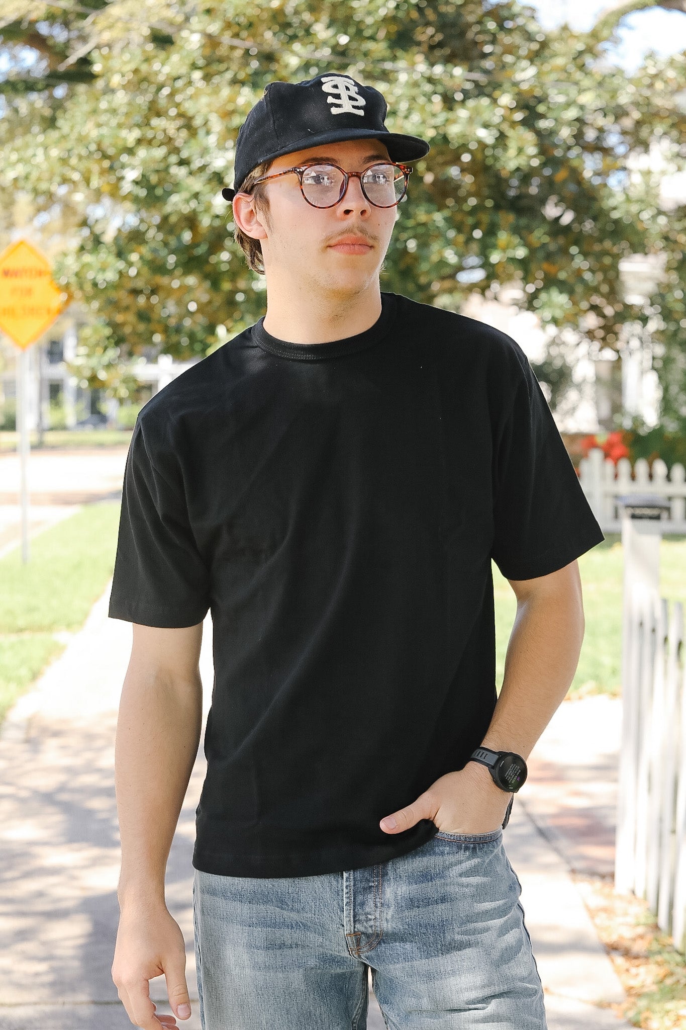 Person wearing a black t-shirt, cap, and glasses outdoors with trees and a street sign in the background.