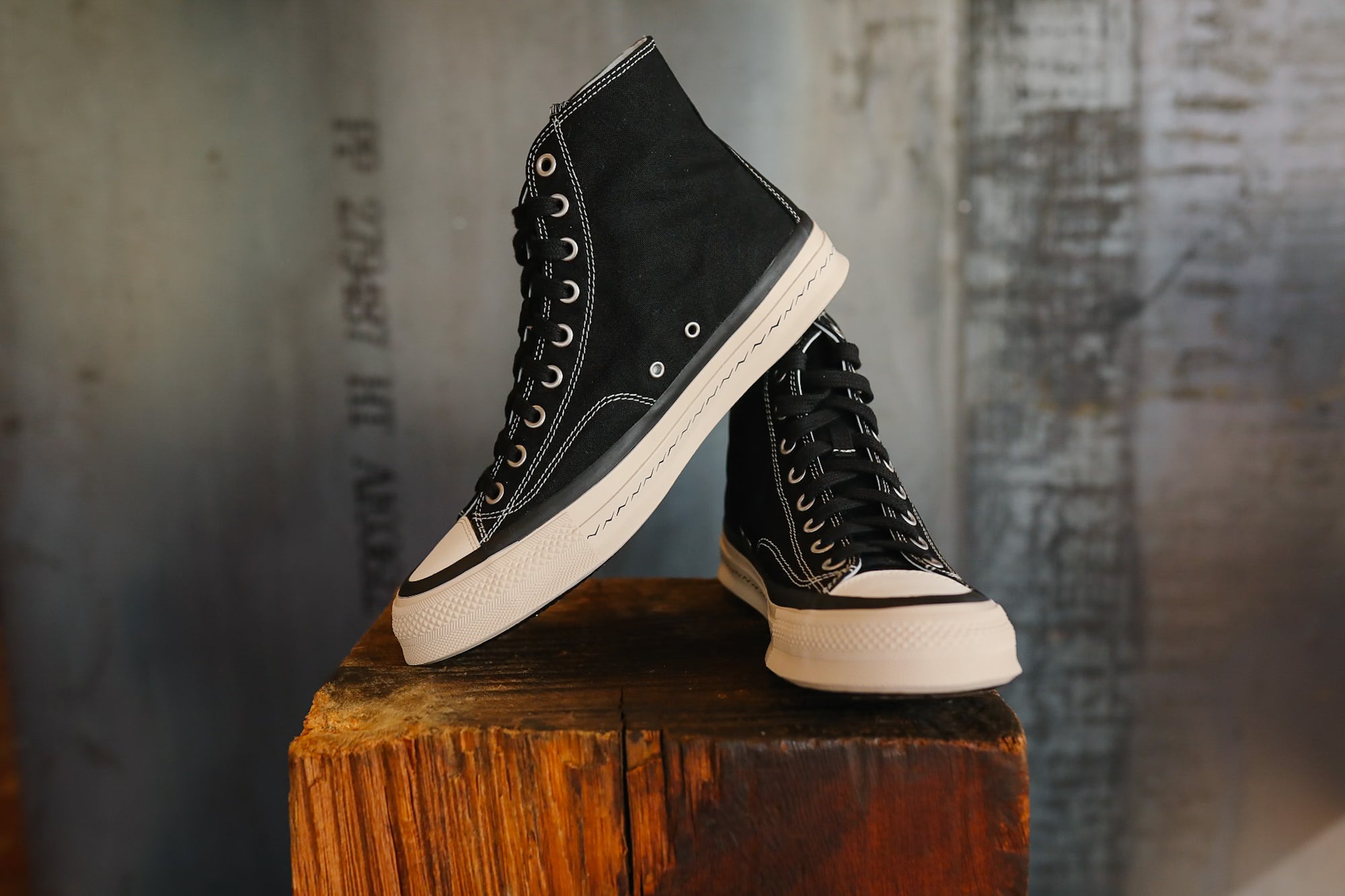 Black high-top sneakers with white soles on a wooden block against a textured wall.