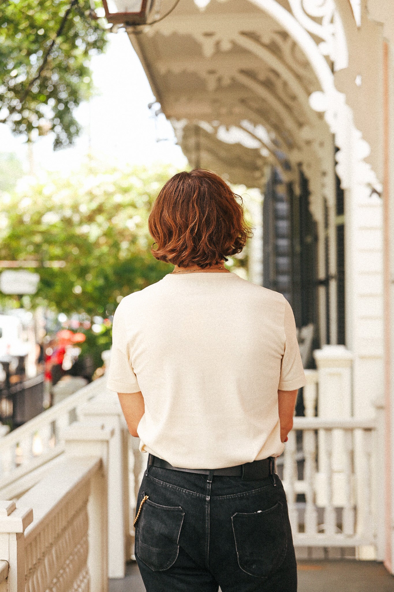 A person viewed from behind wearing a cream short-sleeve t-shirt and dark jeans, standing on a white porch with a street and greenery in the background.