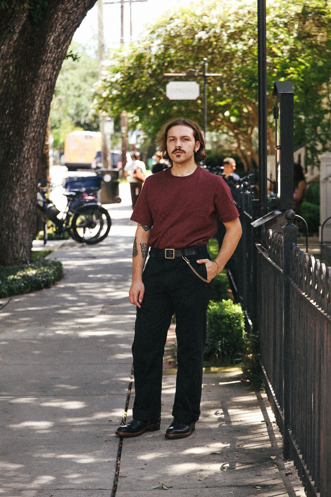 A man stands on a sunny sidewalk wearing a maroon short-sleeve crewneck t-shirt, black pants, and black leather shoes, with his hands in his pockets.