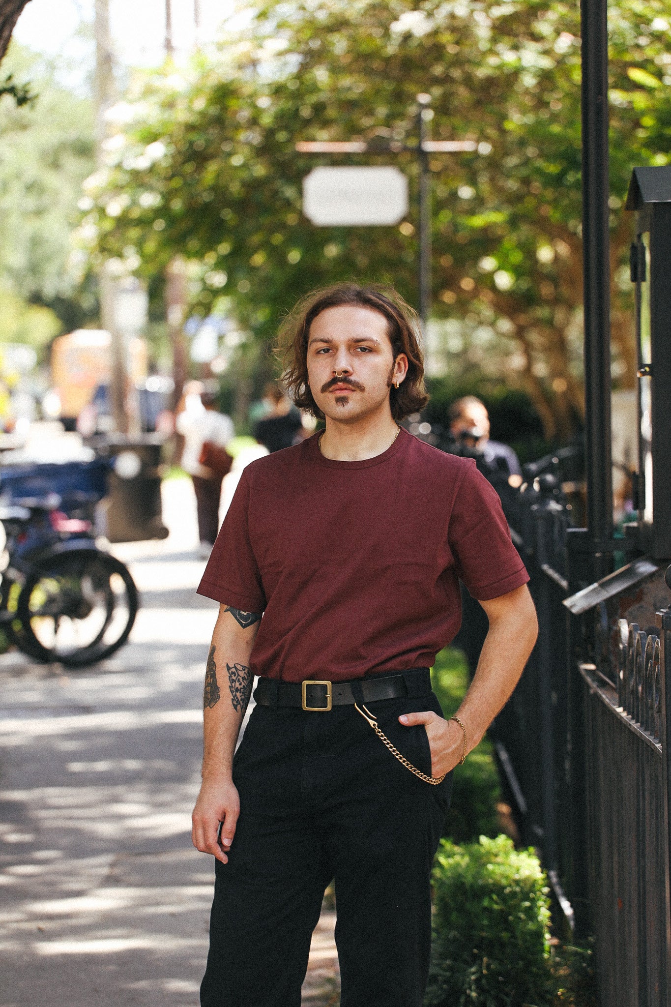 A man with a mustache and shoulder-length hair stands on a sunlit city sidewalk wearing a dark maroon short-sleeve crewneck t-shirt, black trousers, and a black belt with a gold buckle.
