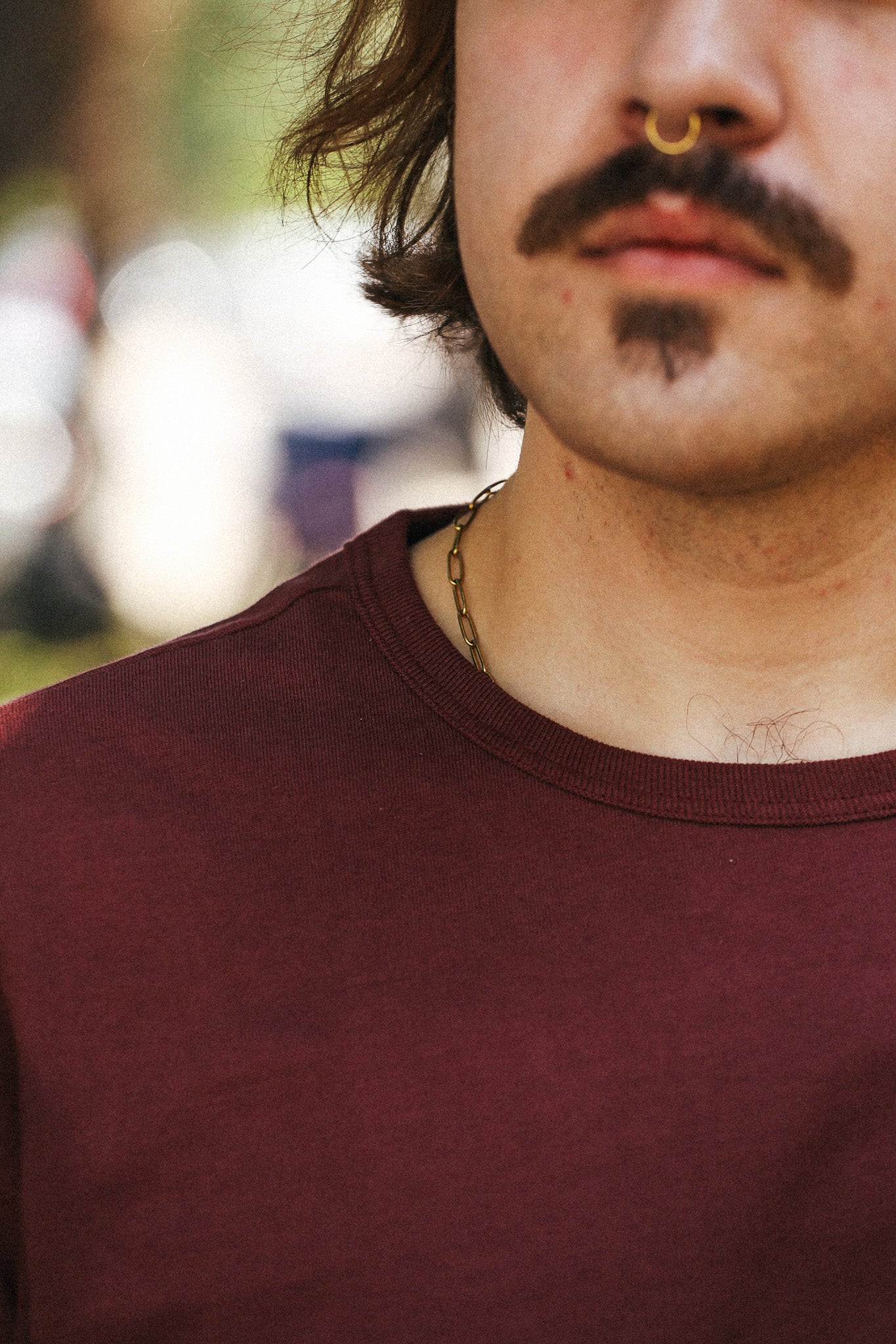 Close-up of a person wearing a burgundy crewneck t-shirt, with a gold chain necklace, nose ring, and gothic mustache and chin tattoo visible against a blurred outdoor background.