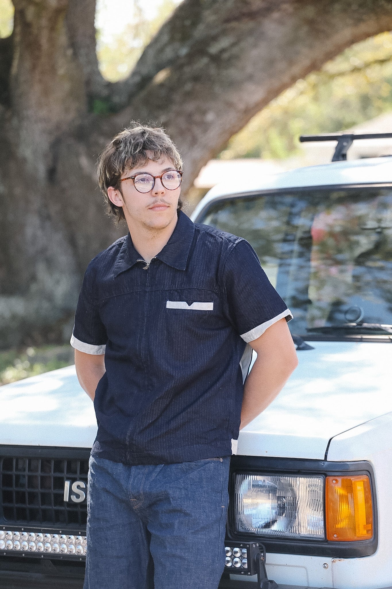 Person wearing glasses and a dark polo shirt standing next to a white vehicle with a tree in the background