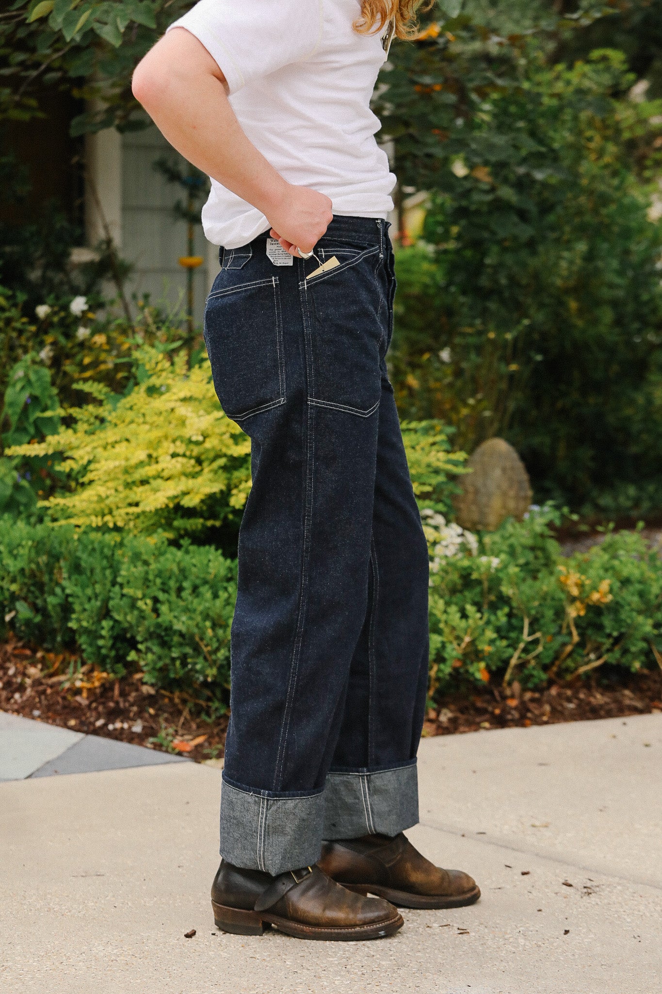 Person wearing dark blue jeans and brown shoes standing outdoors with greenery in the background