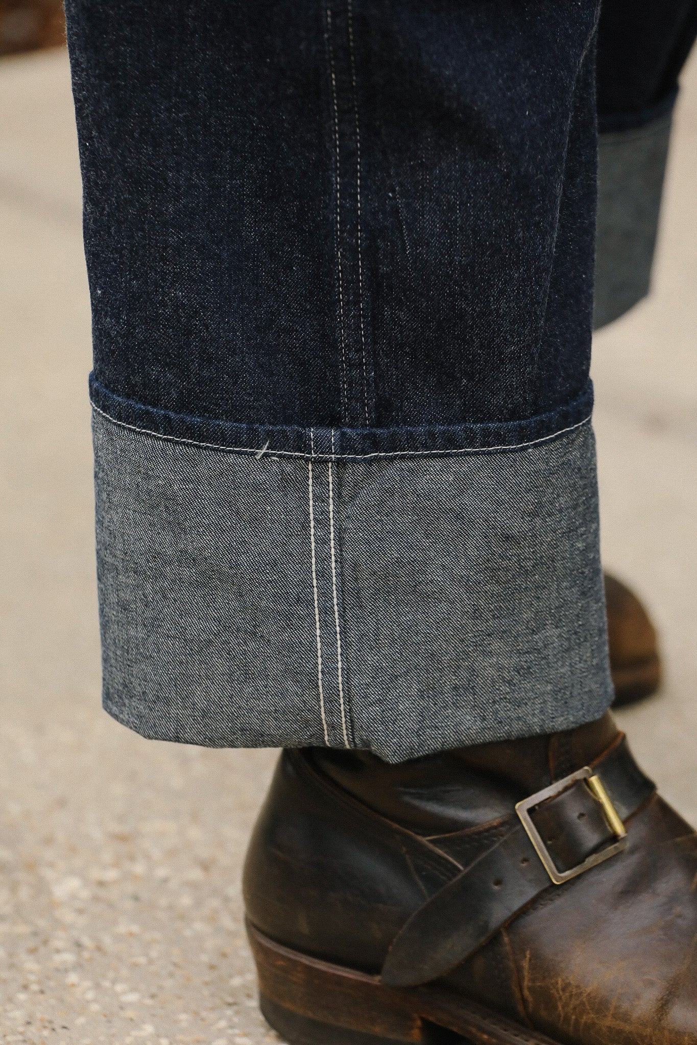 Close-up of dark blue jeans with a gray cuff worn with brown leather shoes on a light background.
