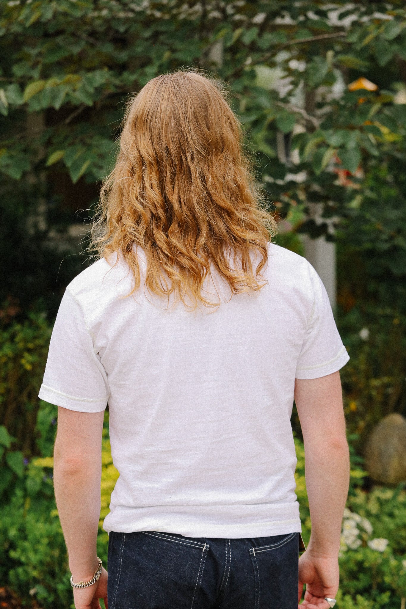 Person with long hair wearing a white t-shirt in a garden setting