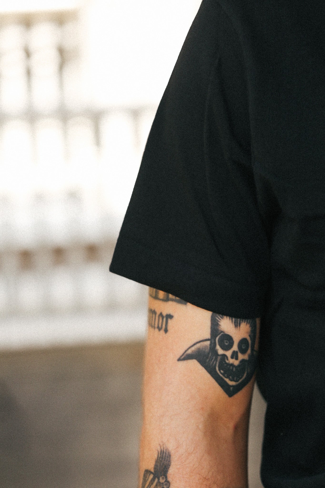 Close-up of a person's forearm wearing a black short-sleeve shirt, with visible black tattoos including a skull design on the upper arm.