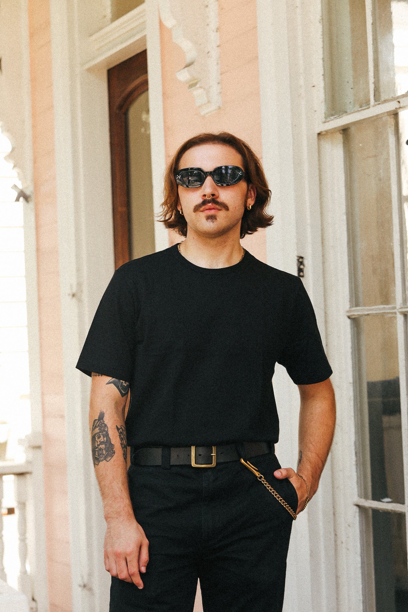 A man with a mustache and sunglasses wearing a black crewneck t-shirt and black pants, standing on a porch in front of a peach-colored building.