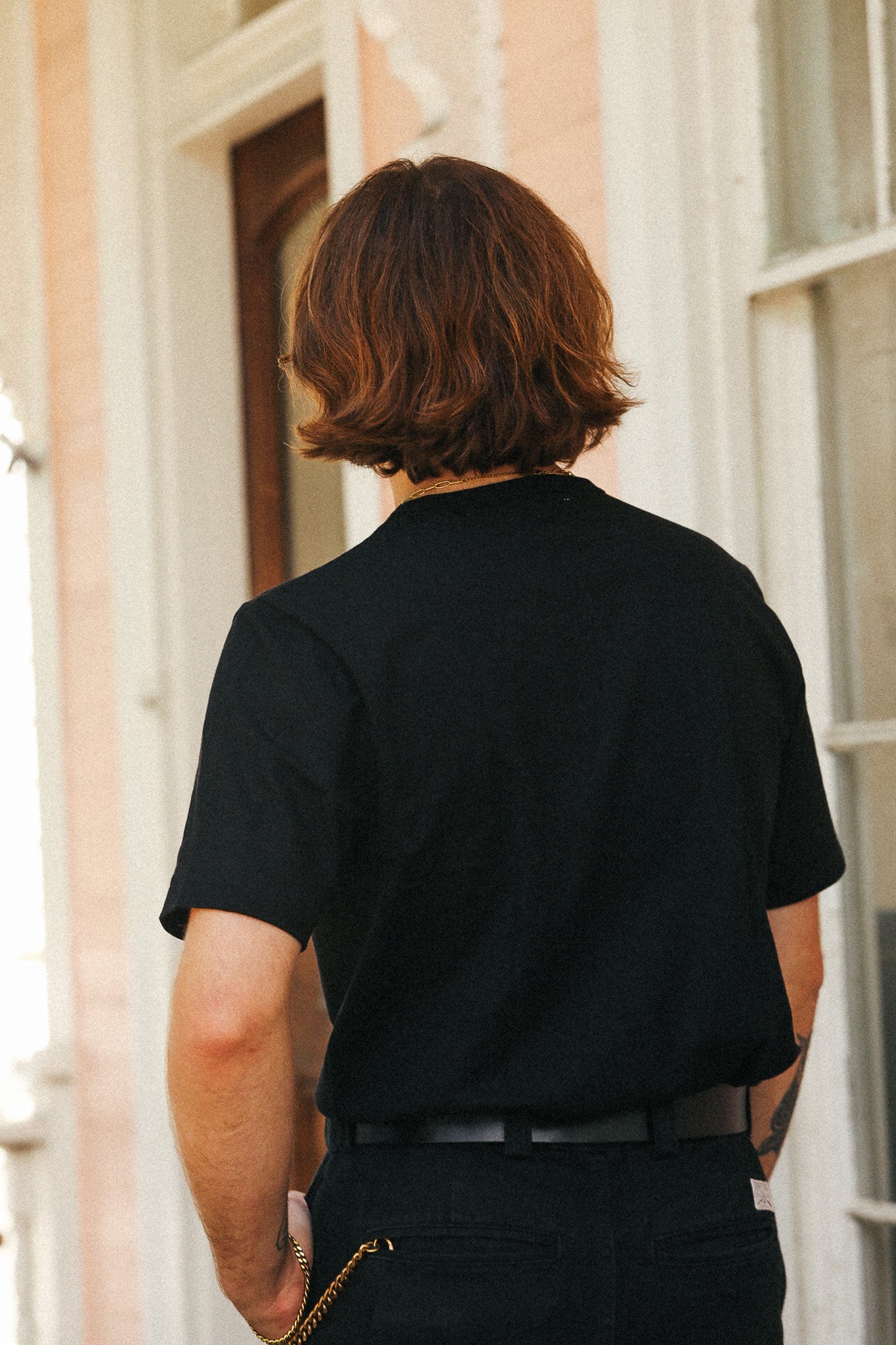 A person viewed from behind wearing a black short-sleeve t-shirt and black pants, standing outdoors in front of a light-colored building.