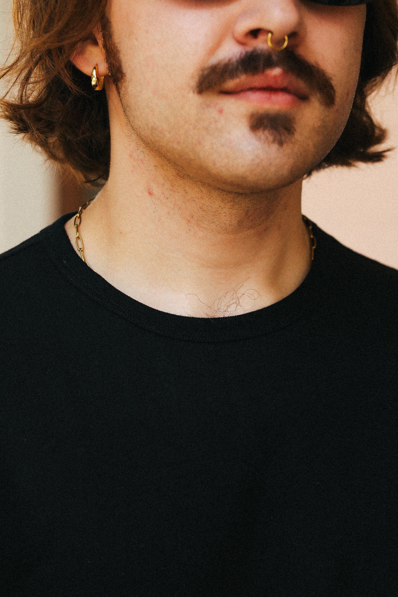 Close-up of a person wearing a black crew-neck t-shirt, a gold hoop earring, a nose ring, and a gothic mustache.