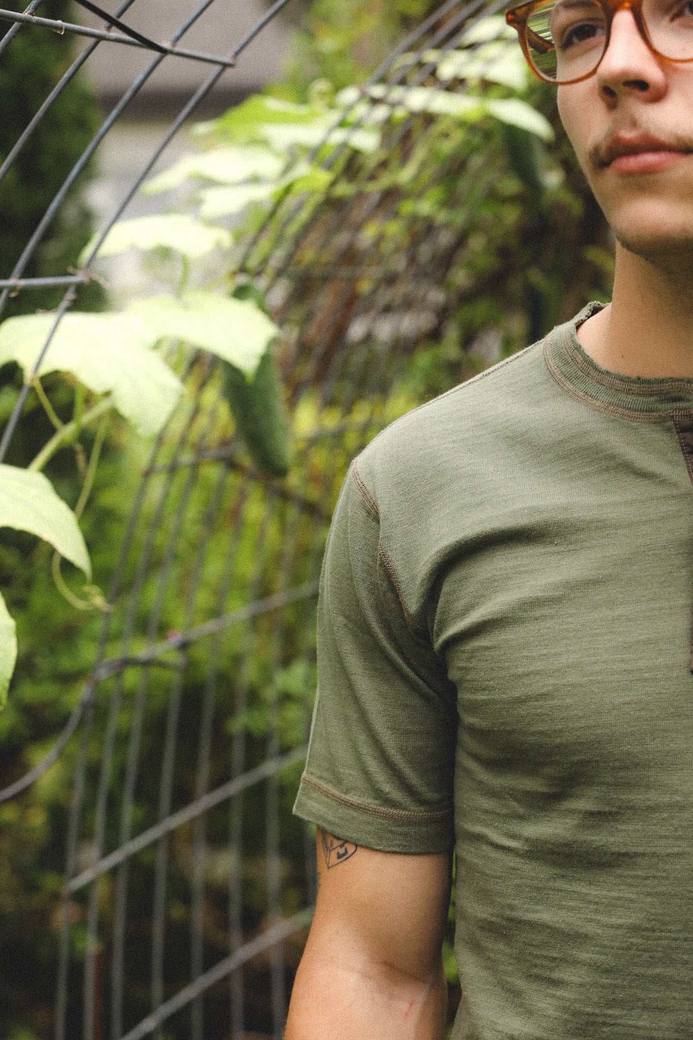 Person wearing a green t-shirt standing in front of a wire fence with plants.
