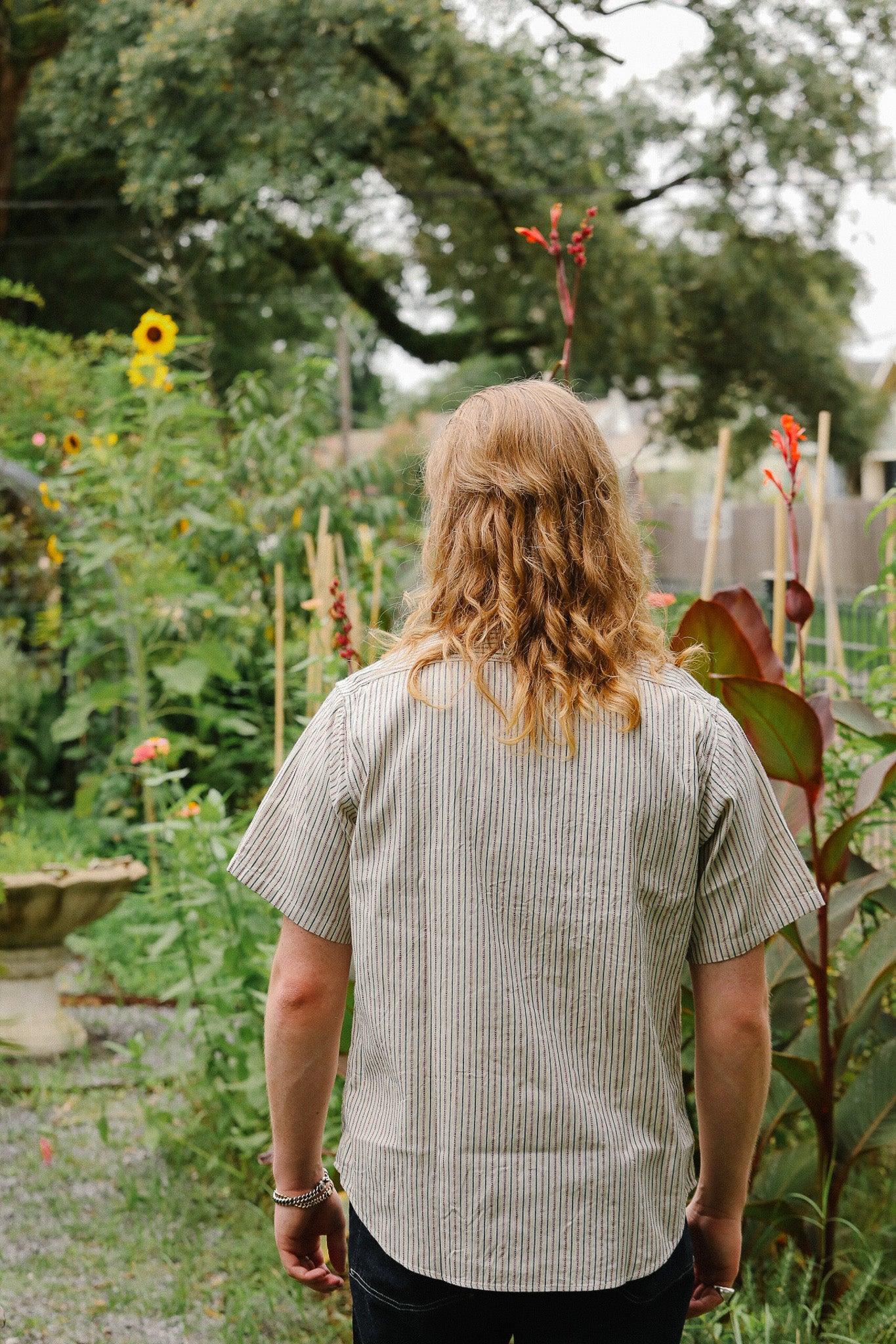 Person with long hair standing in a garden with various plants and flowers.