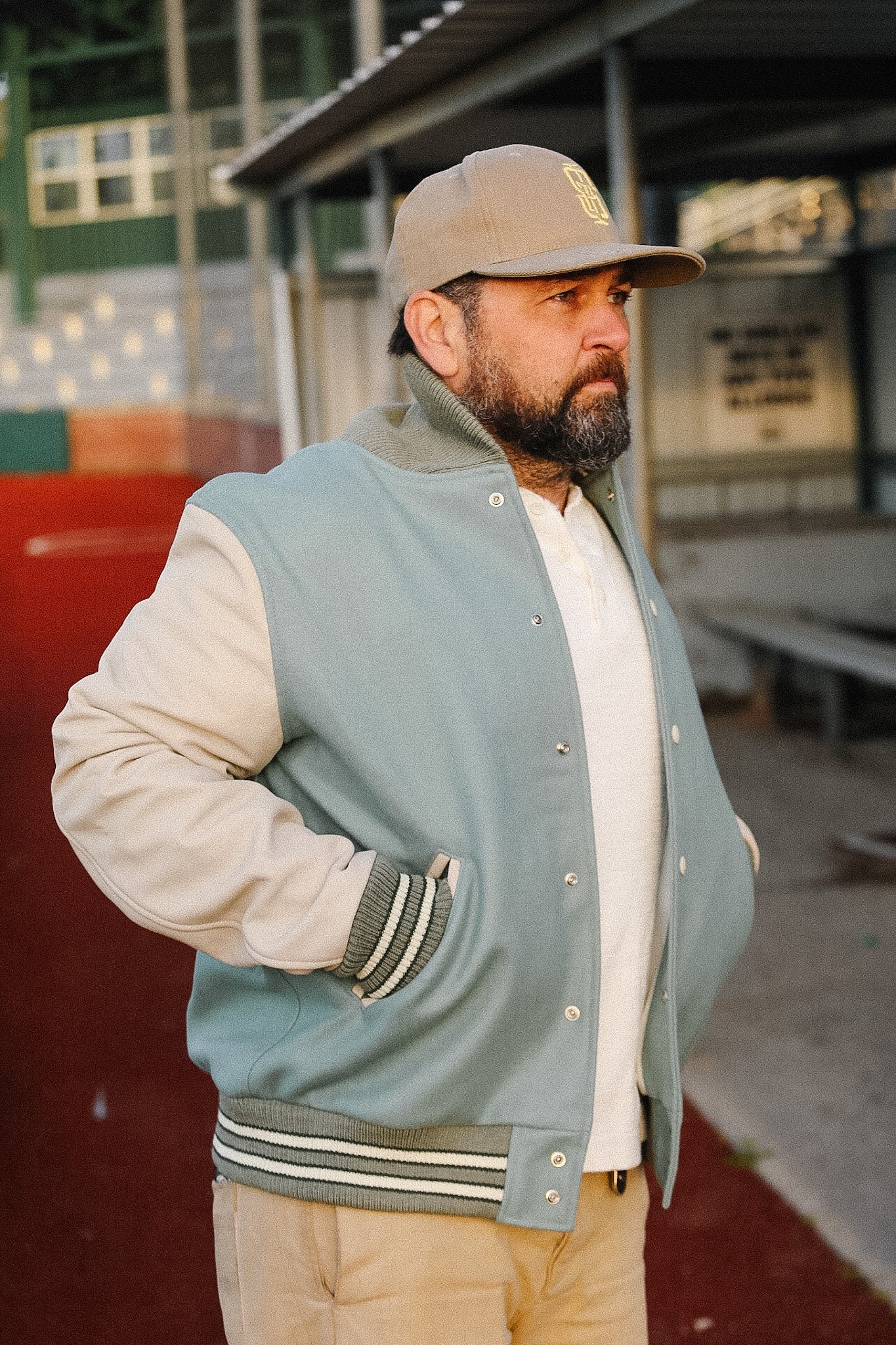 Man wearing a light blue varsity jacket with beige sleeves and a cap, standing in an outdoor setting.