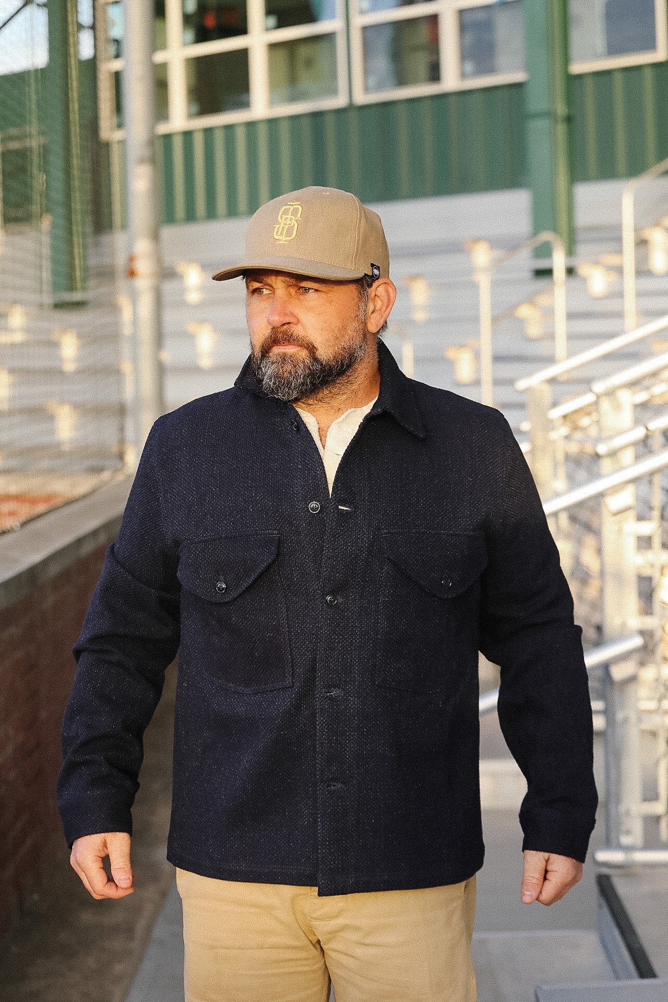 Man wearing a dark jacket and beige cap standing on a rooftop with metal railings and green wall in the background.