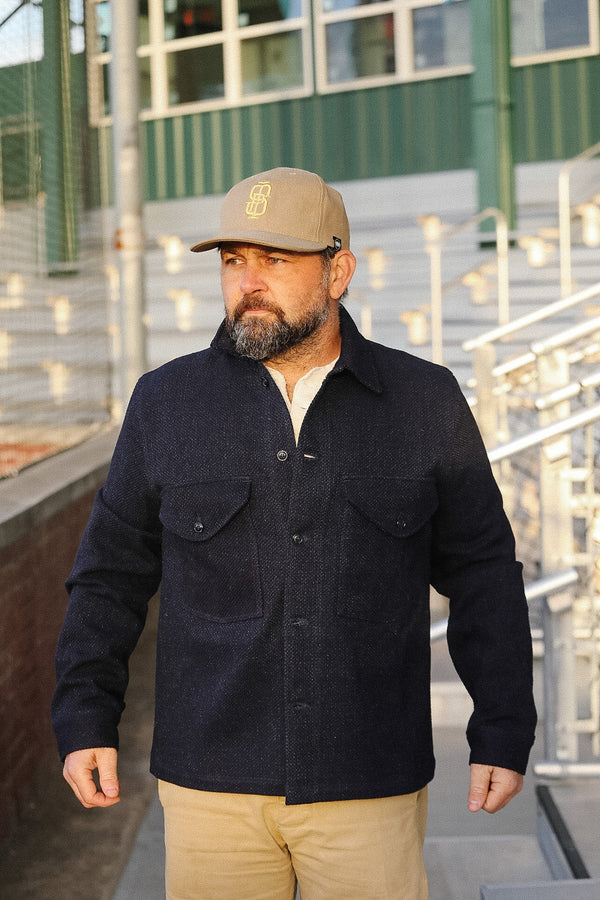 Man wearing a dark jacket and beige cap standing on a rooftop with metal railings and green wall in the background.