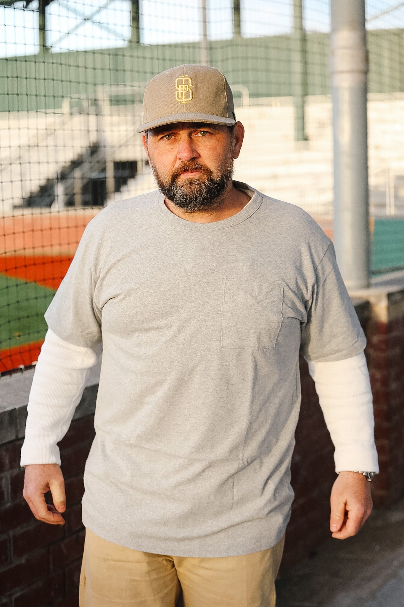Man wearing a gray long-sleeve shirt and beige cap with a logo, standing outdoors.