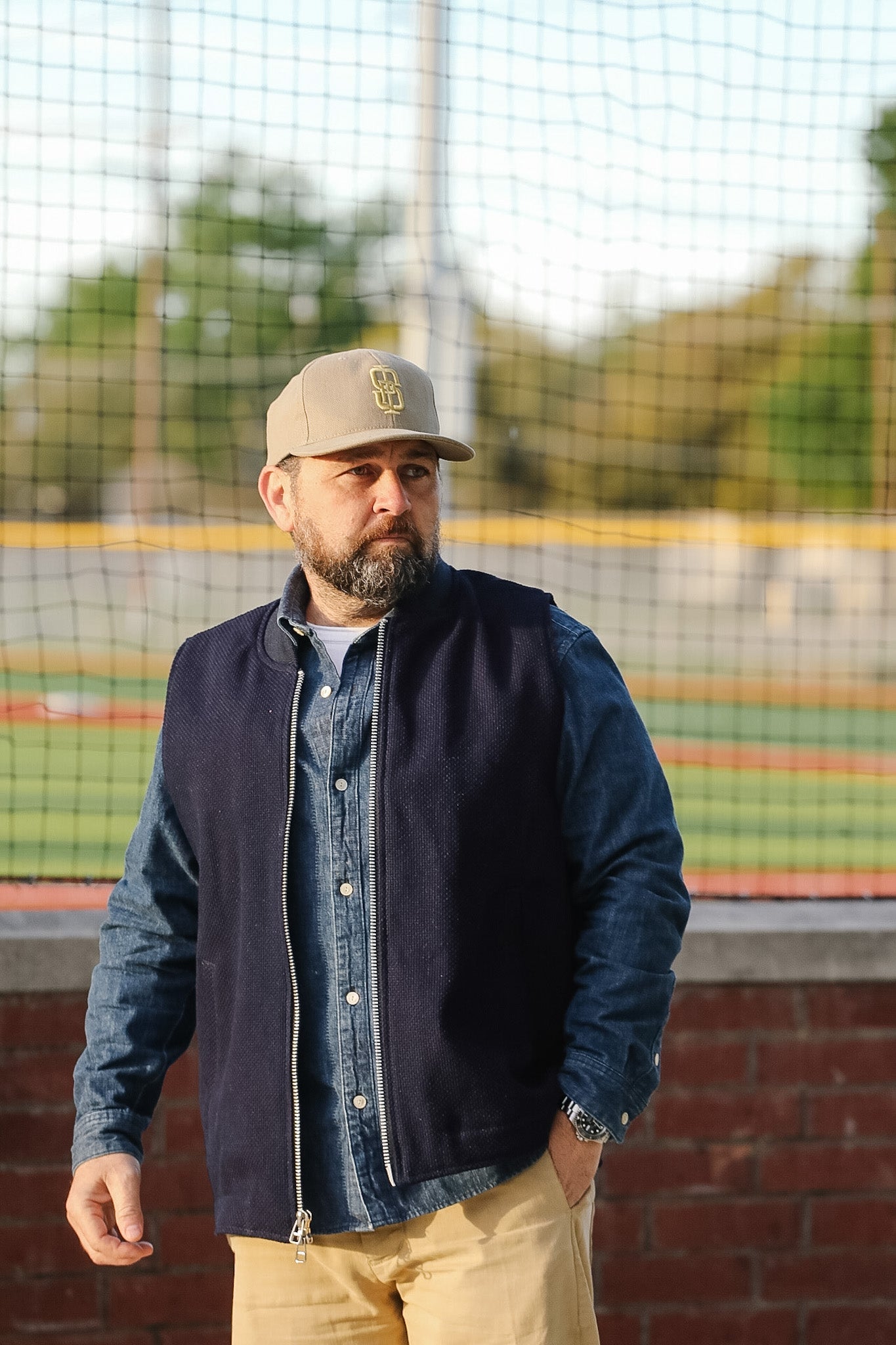 Man wearing a navy jacket, denim shirt, and beige cap standing on a sports field.