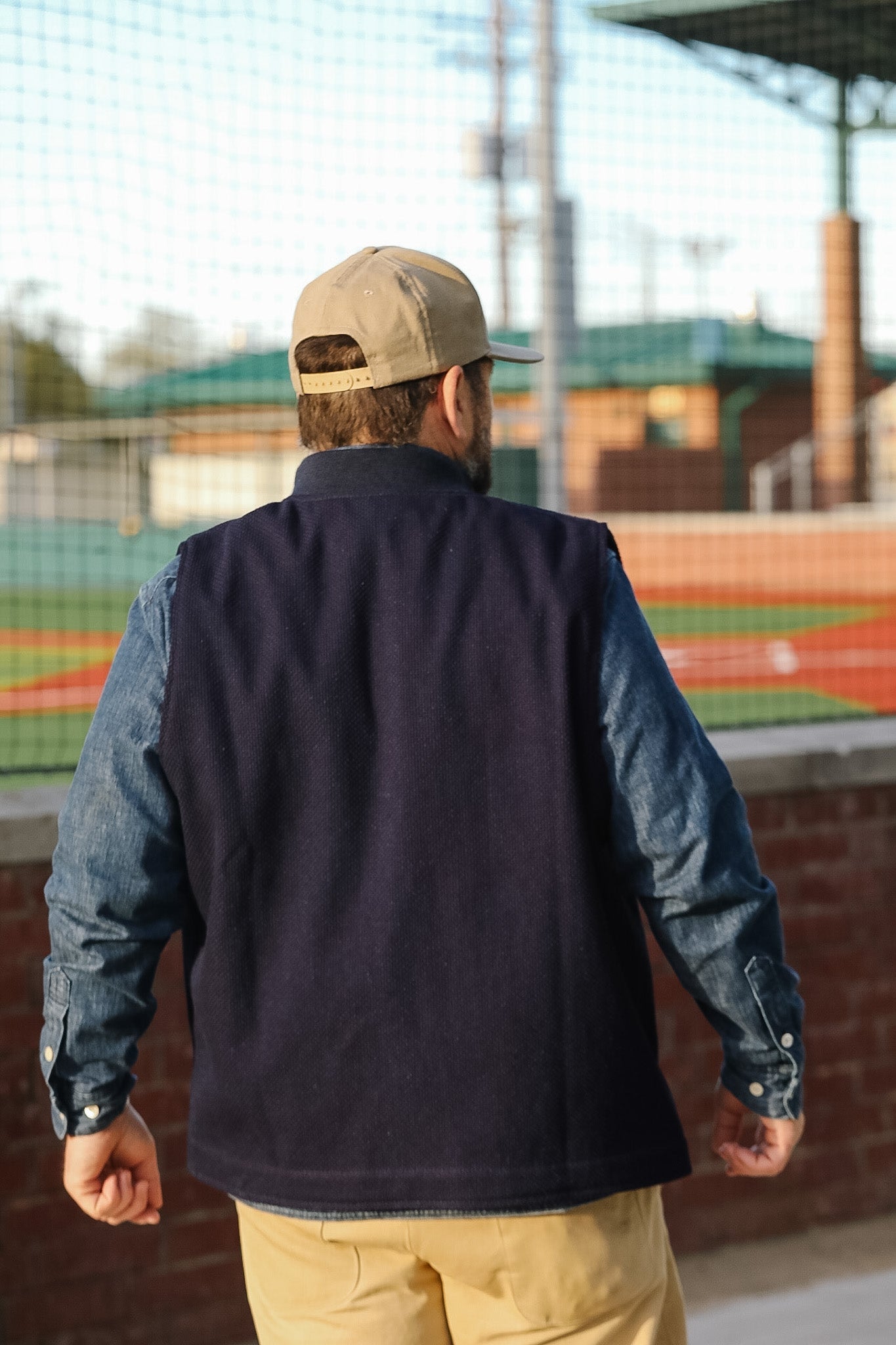 Man wearing a navy vest over a denim shirt with beige pants and cap, standing in front of a sports field.