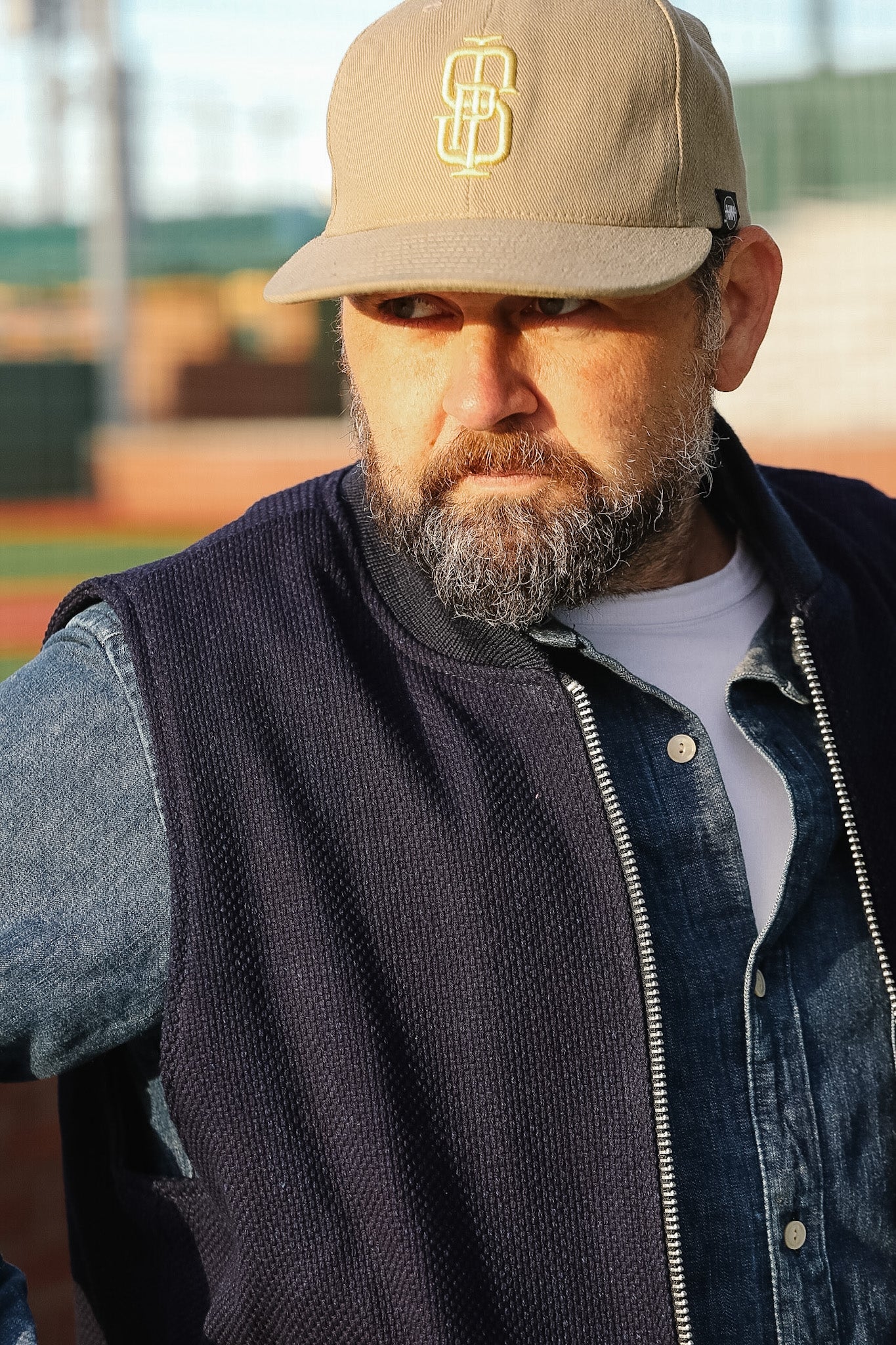 Man wearing a beige cap with a logo, dark vest, and denim jacket on a sports field.