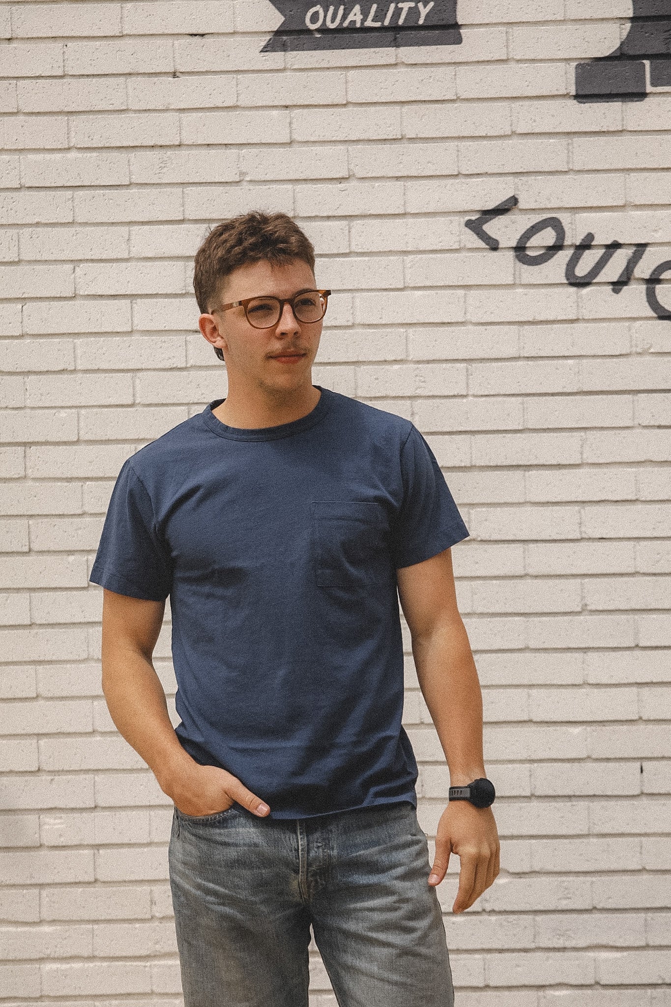 Man wearing a navy blue t-shirt and glasses standing against a textured wall.
