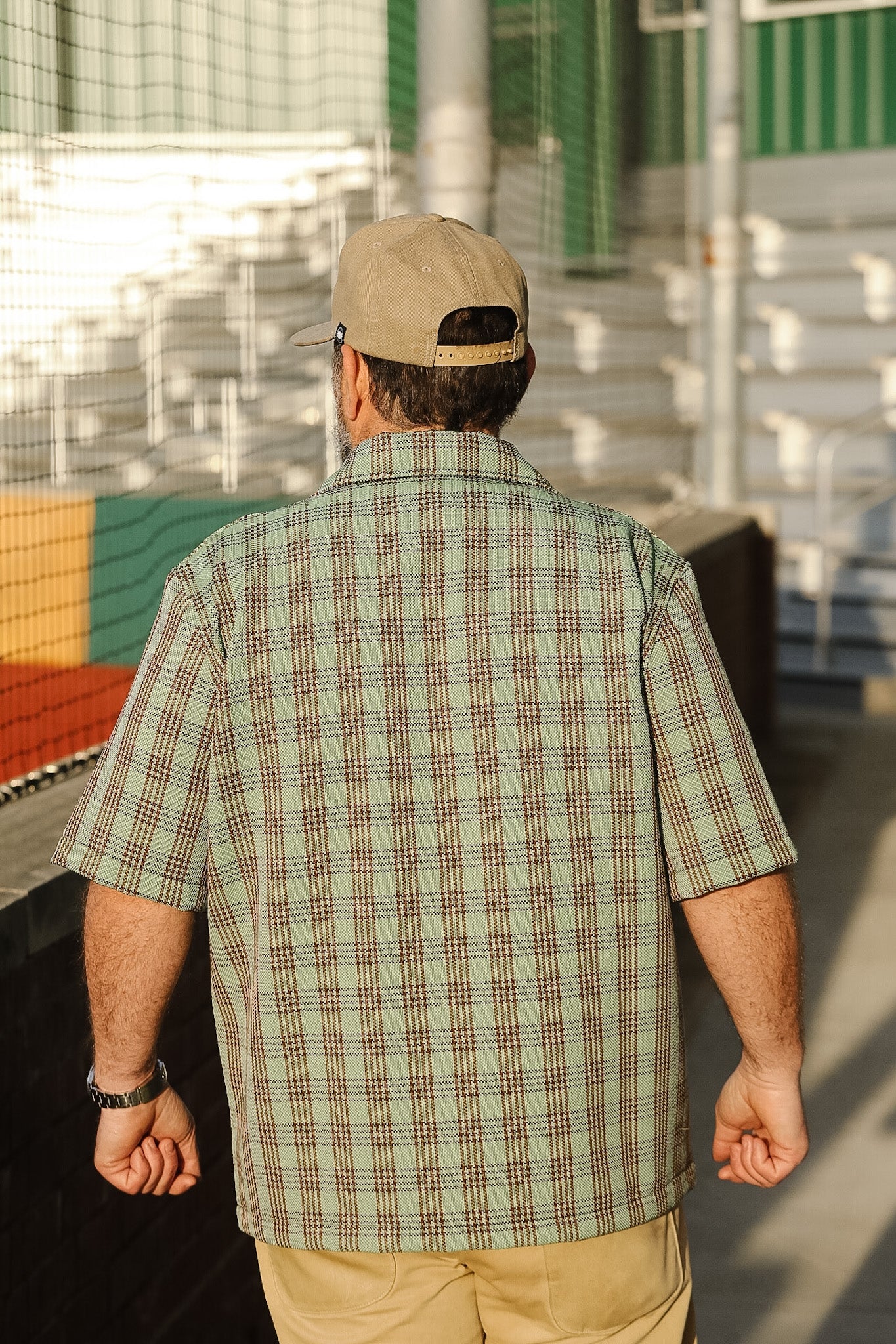 Man wearing a plaid shirt and cap walking away from the camera with industrial containers in the background.