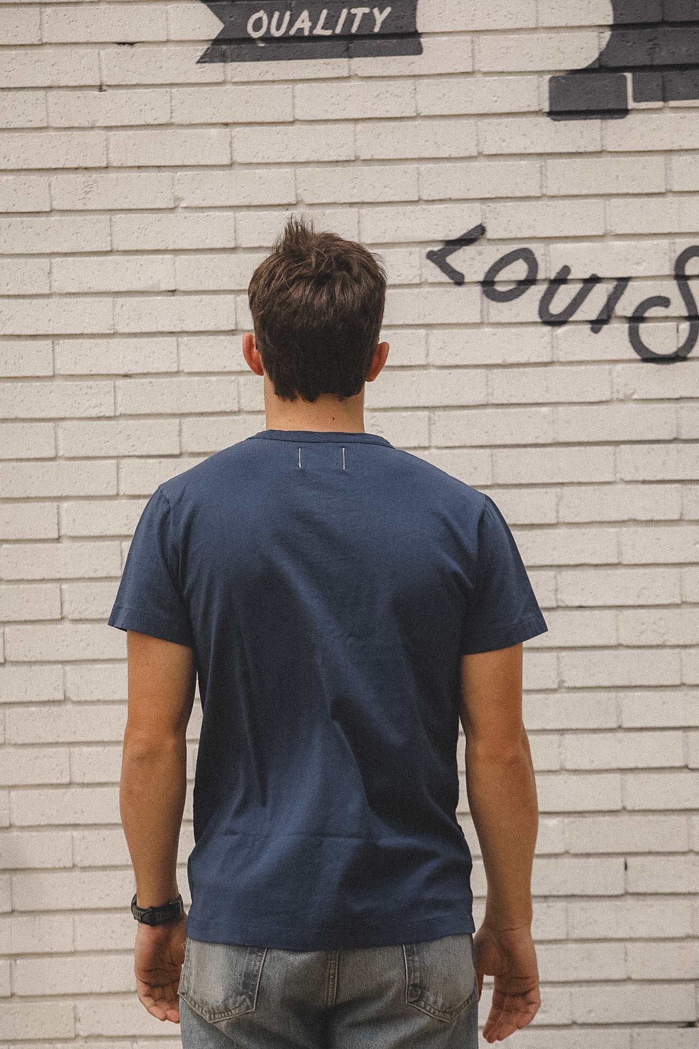Person wearing a navy blue t-shirt standing in front of a textured wall with graffiti.