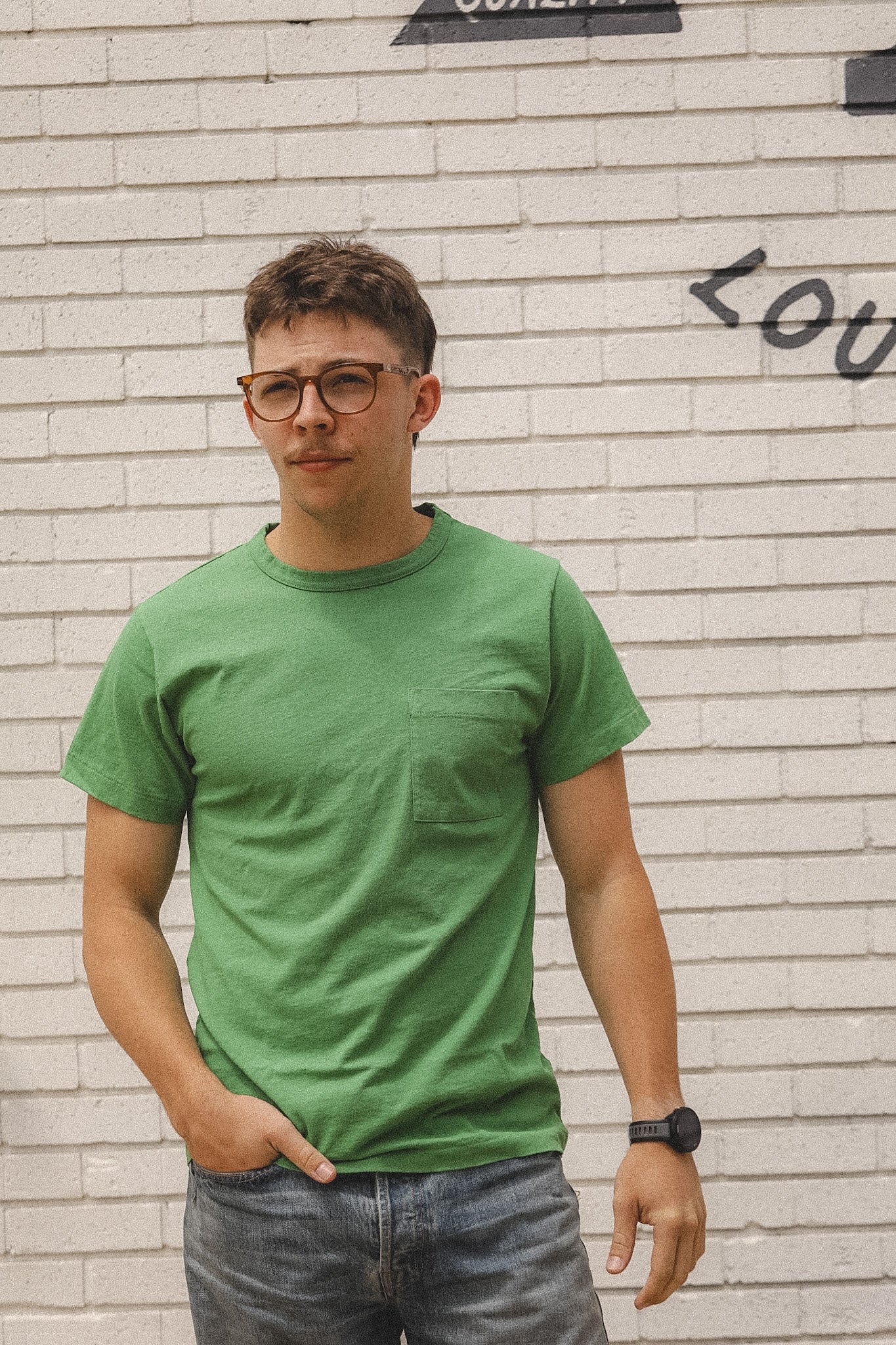 Man wearing a green t-shirt and glasses standing against a light brick wall.