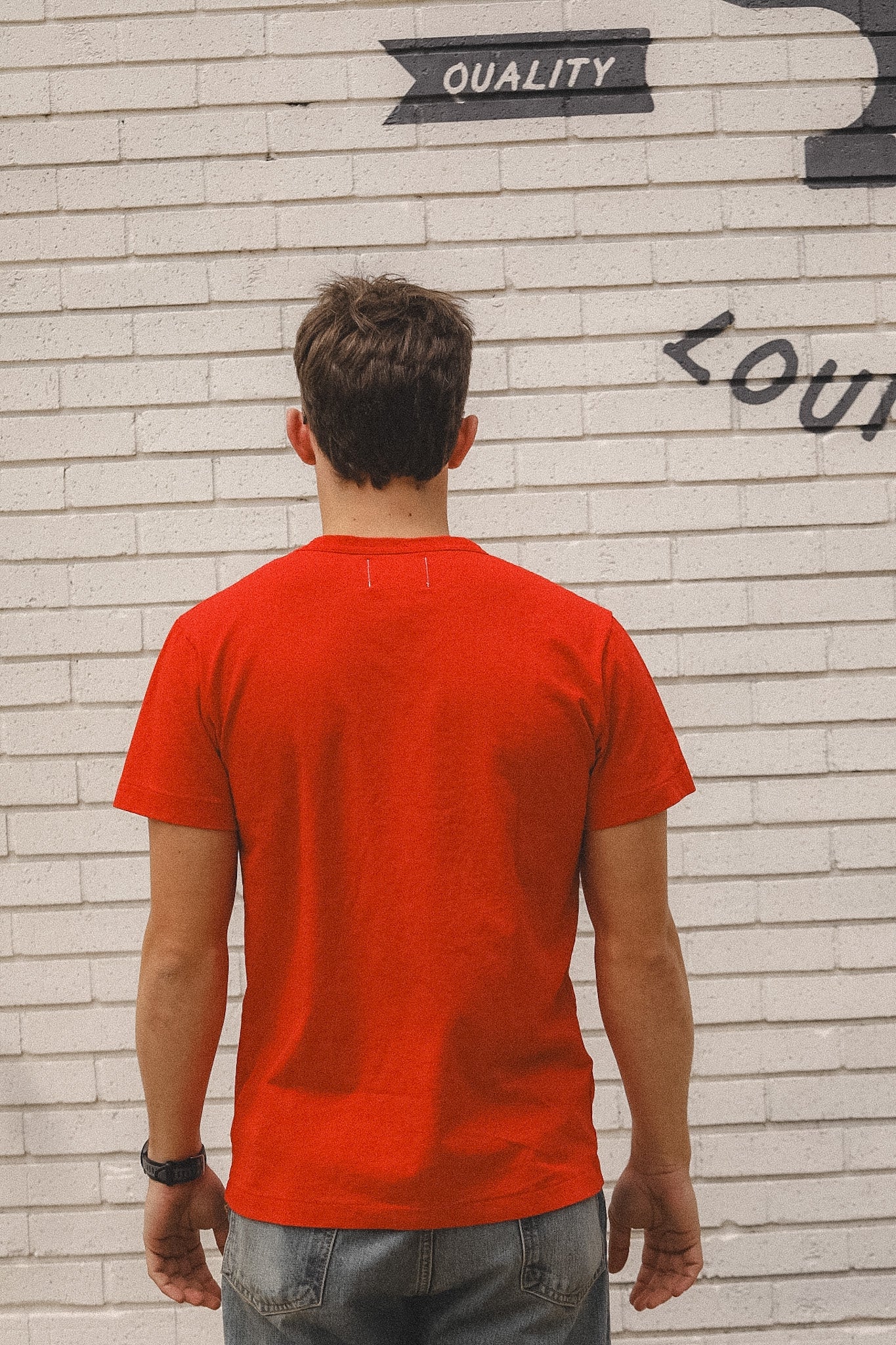 Person wearing a red t-shirt standing against a textured wall with graffiti.