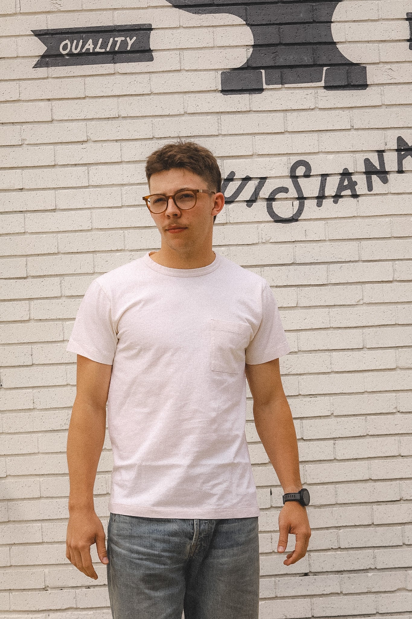 Man wearing a white t-shirt and glasses standing in front of a brick wall with graffiti.